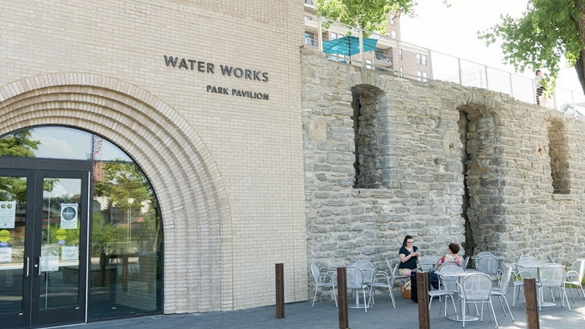Outdoor seating area with metal chairs and tables beside a brick and stone building labeled "WATER WORKS PARK PAVILION," two people sit and chat near the stone wall under trees.