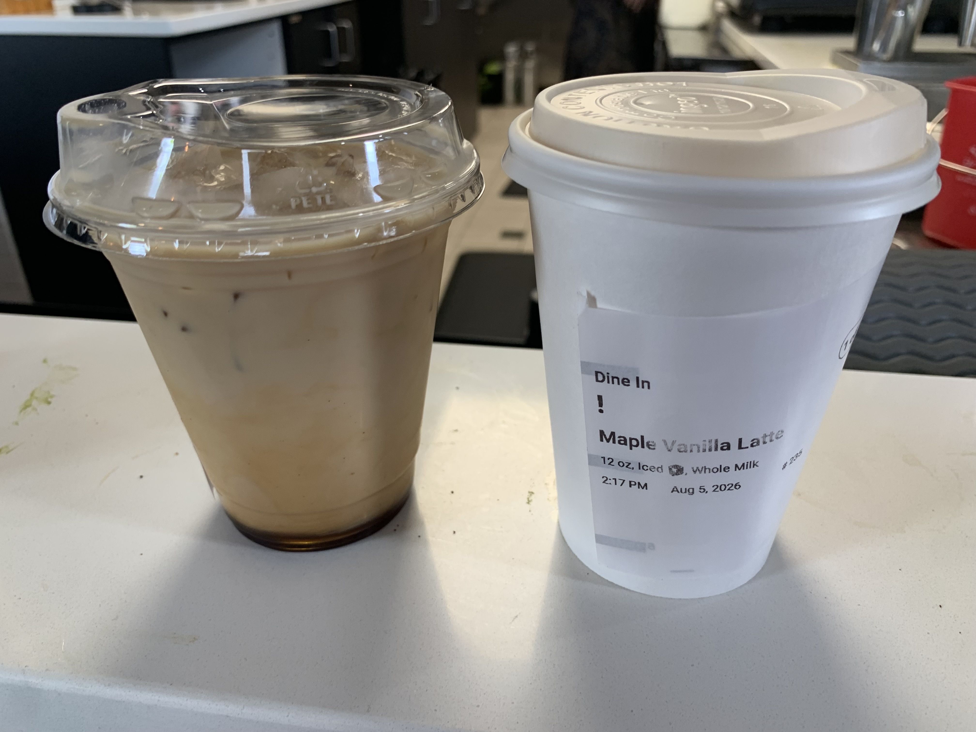 Two coffee cups on a white counter: an iced coffee in a clear dome lid on the left and a hot beverage in a white takeout cup with lid and sleeve on the right, in a cafe setting.