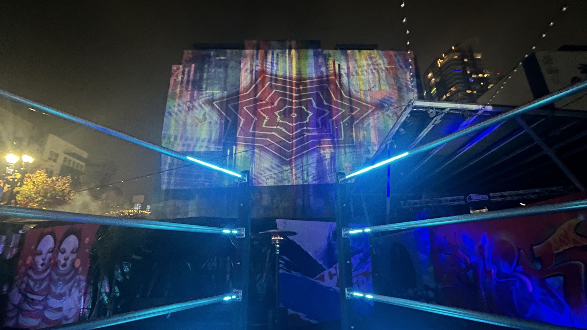 Colorful lights are projected on a city building with a blue LED sculpture as part of a digital art installation. 