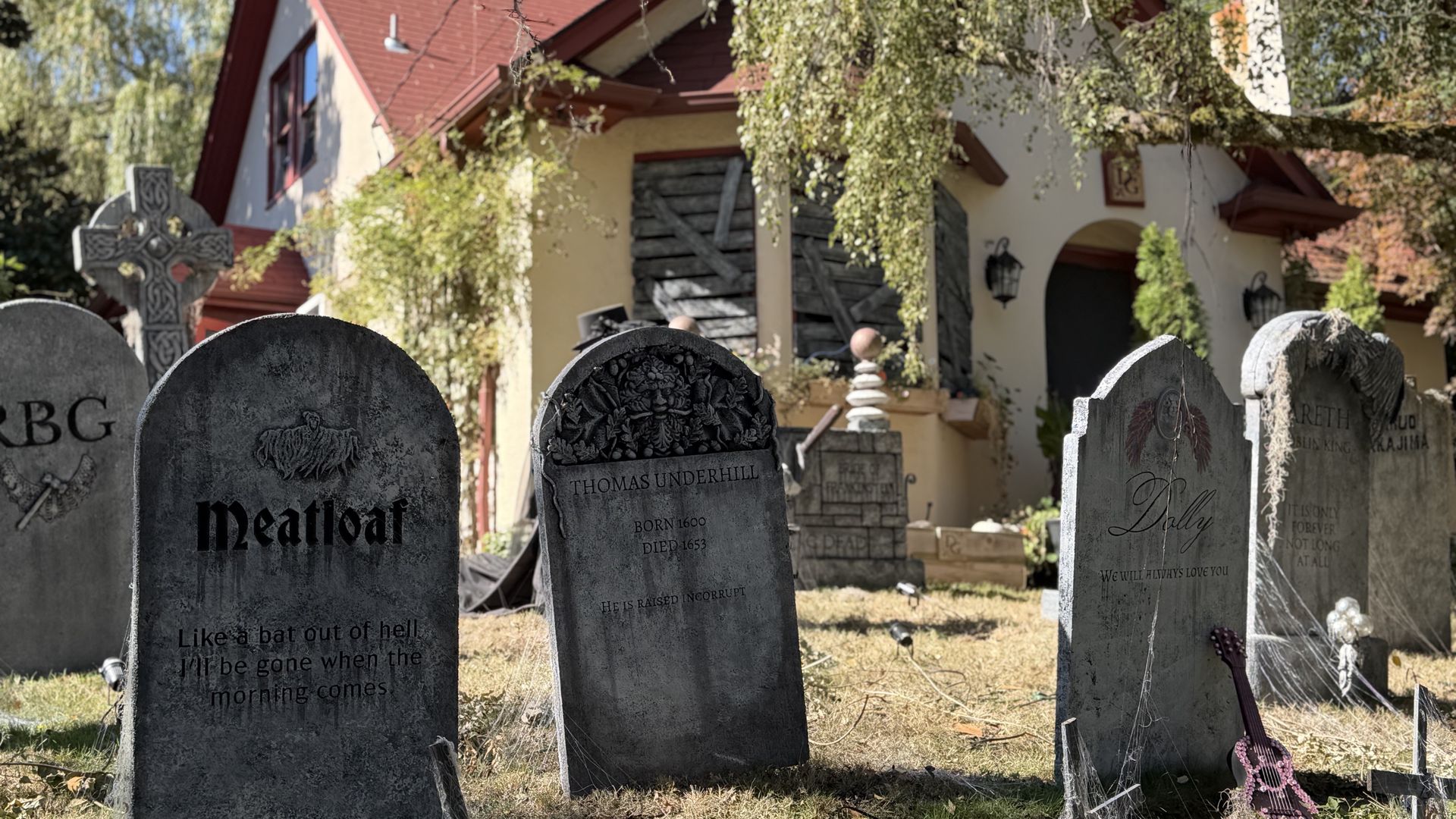 Graveyard in foreground with weathered headstones; one stone bears a large carved name. Behind it, a cream house with a red pitched roof and hanging vines; a guitar rests near the right edge.