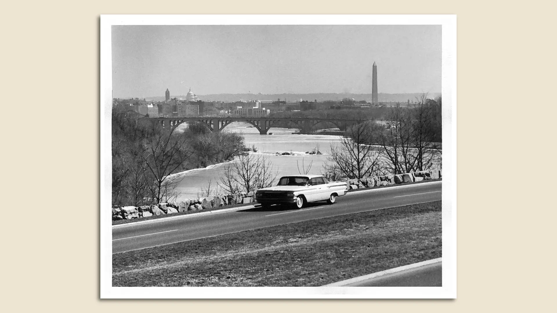 Black-and-white photo of a vintage convertible driving along a road beside a river, leafless trees line the bank, with a distant bridge and Washington, D.C. skyline including the Capitol dome and a tall obelisk.