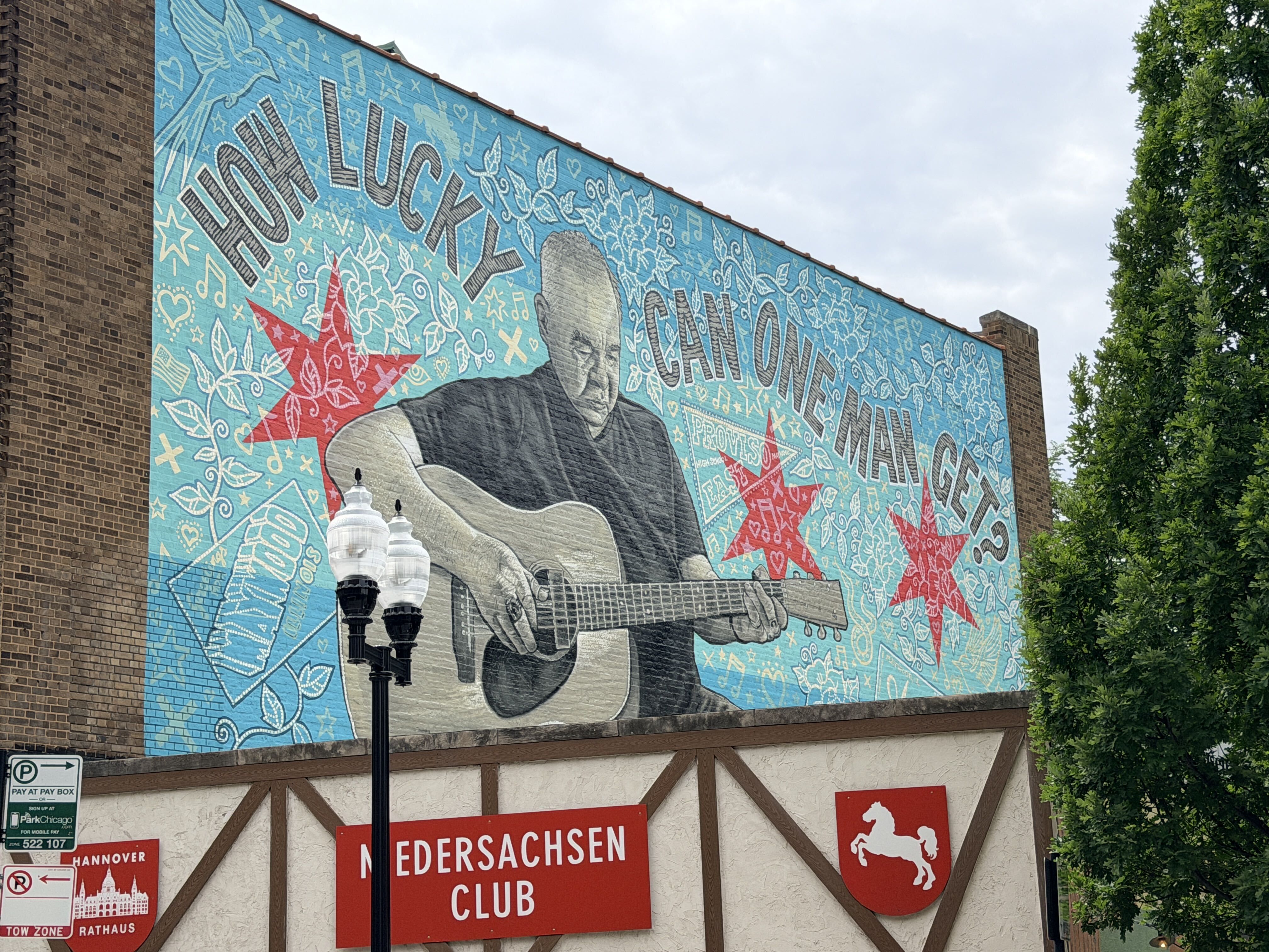 Large mural on a brick wall featuring a grayscale guitarist with a guitar, set against a blue decorative background with stars, leaves, and musical notes; trees to the right and a cloudy sky.