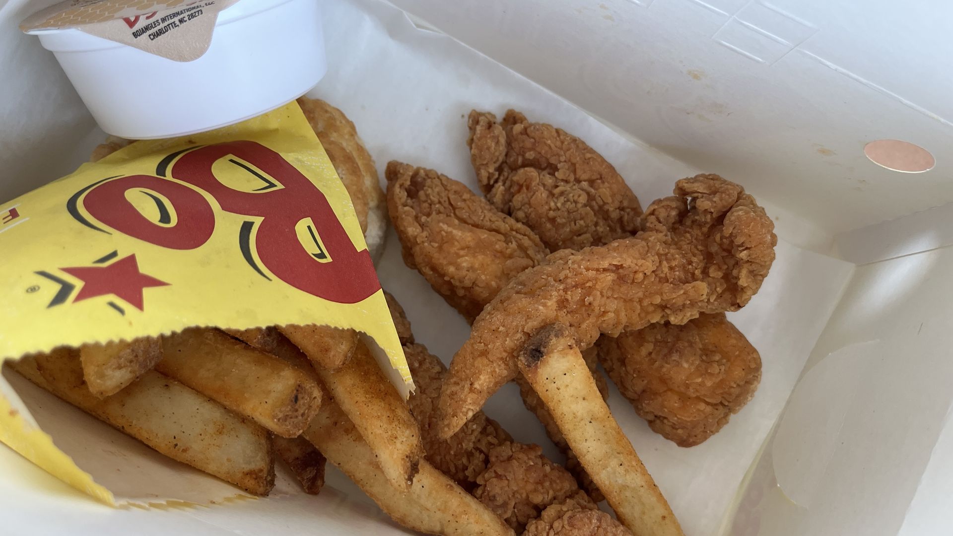 Inside a white takeout box, golden-brown fried chicken pieces and seasoned fries peek from a yellow bag, with a white cup on top, all on white packing paper.