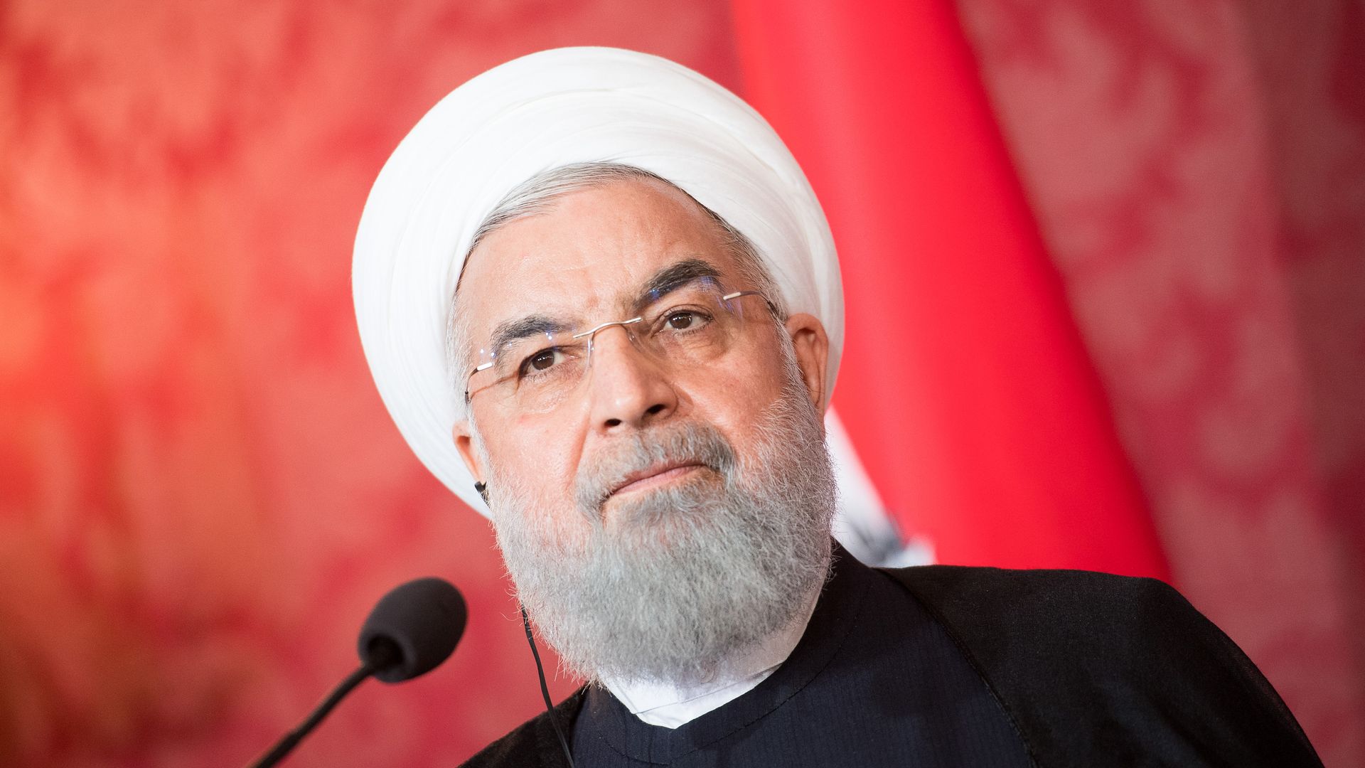 Hassan Rouhani in white head covering, black shirt, before red background looking stern.