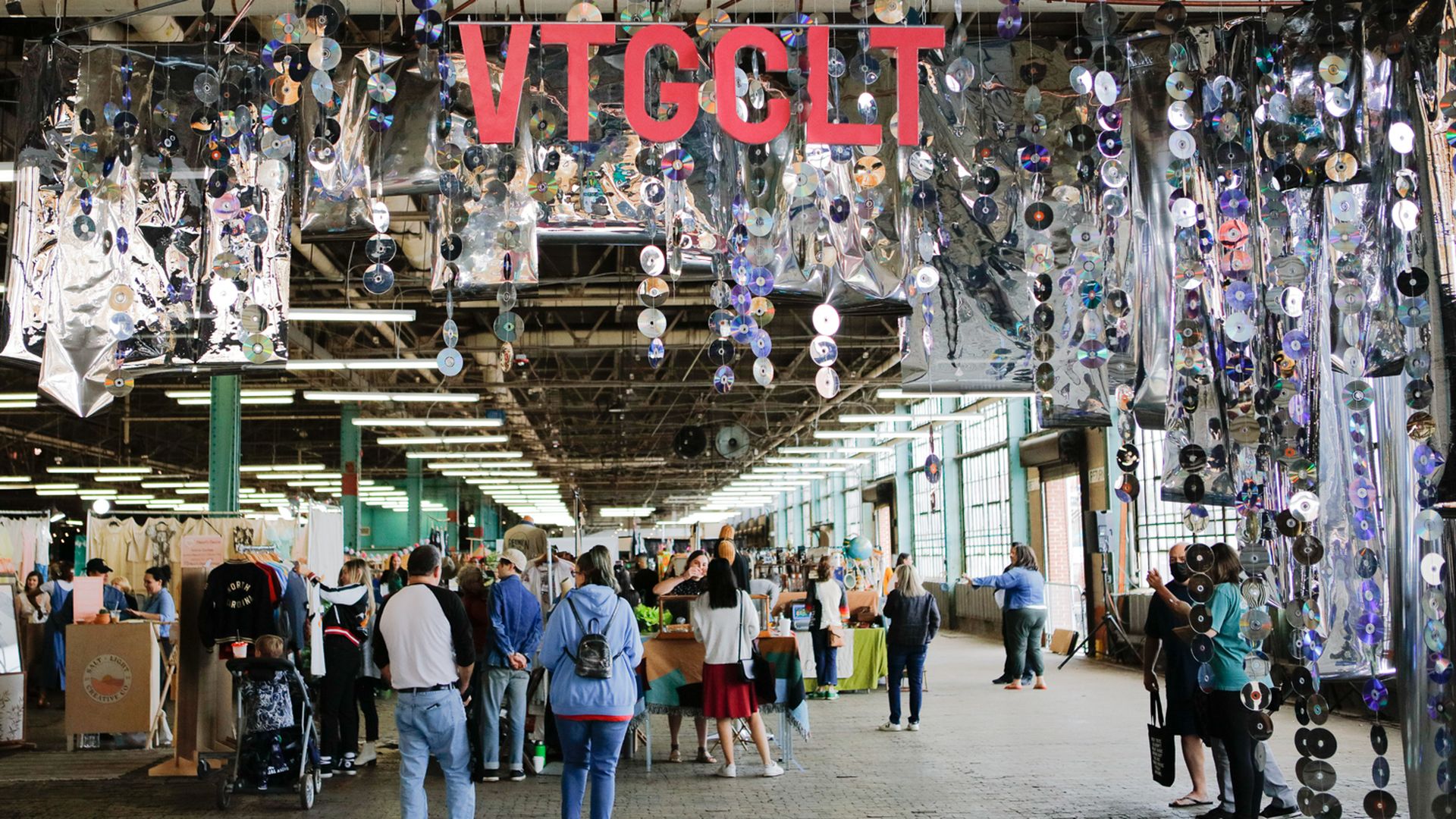 Indoor market with people browsing stalls beneath hanging decorations made of CDs and reflective silver panels, with large red letters spelling VTGCLT overhead.