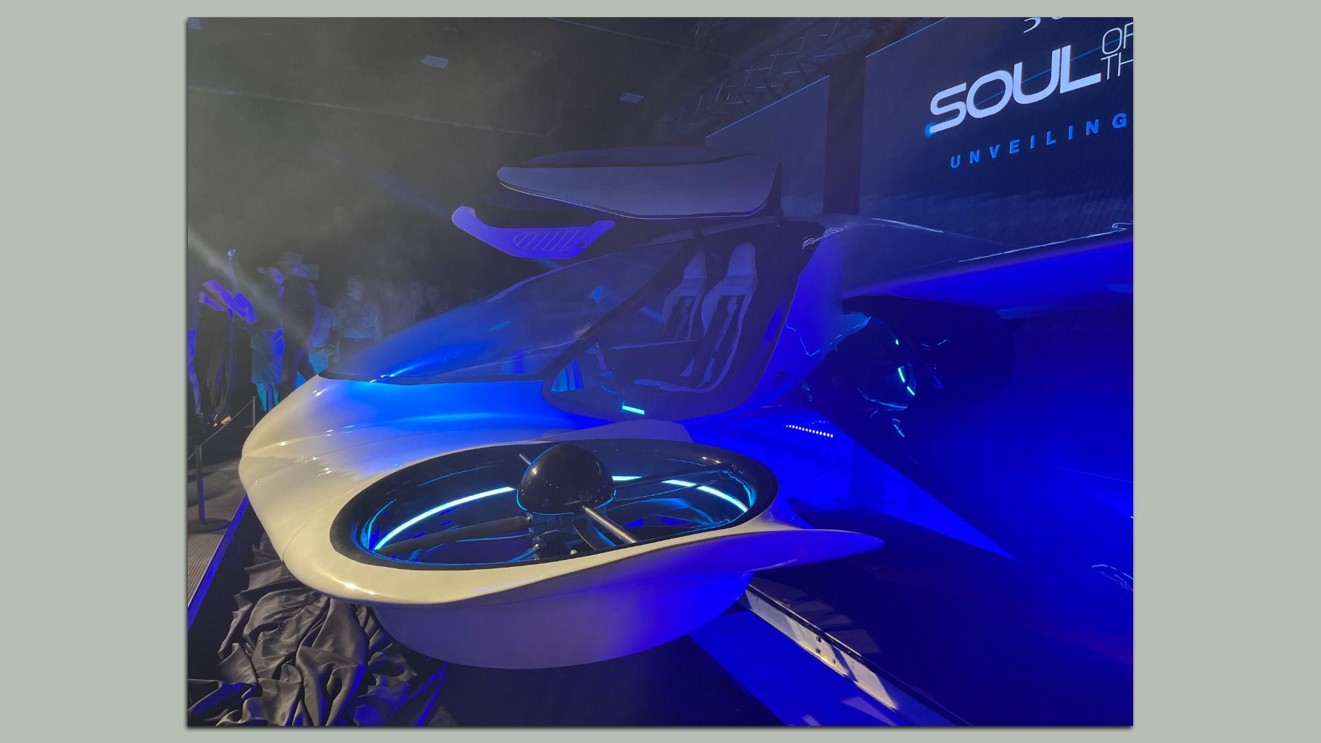 Futuristic white EVTOL aircraft on a dim stage with blue neon lighting; gull-wing doors open, two seats visible inside. A circular blue-lit platform sits in the foreground. 