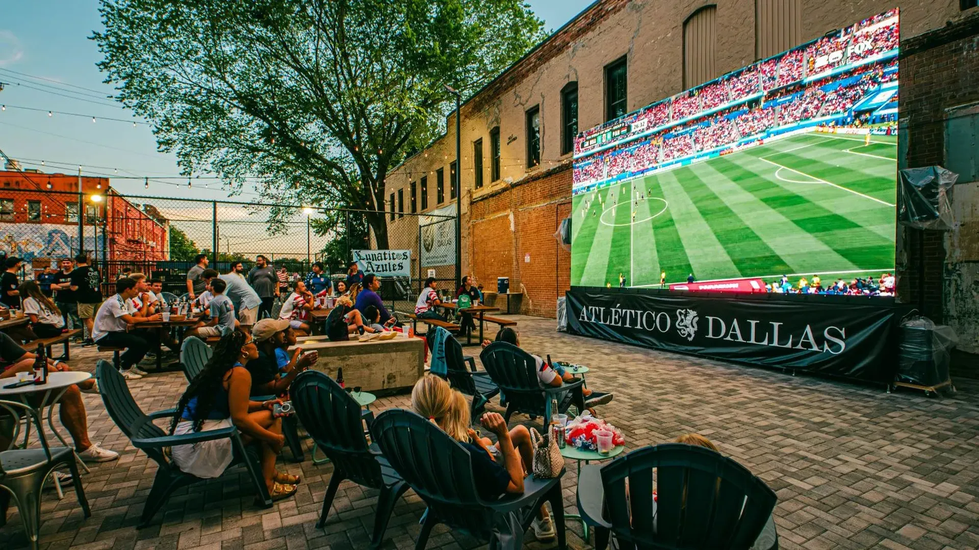 Outdoor urban courtyard with string lights and brick walls, crowd watching a soccer match on a large screen; people sit in Adirondack chairs and picnic tables; banner says ATLÉTICO DALLAS.