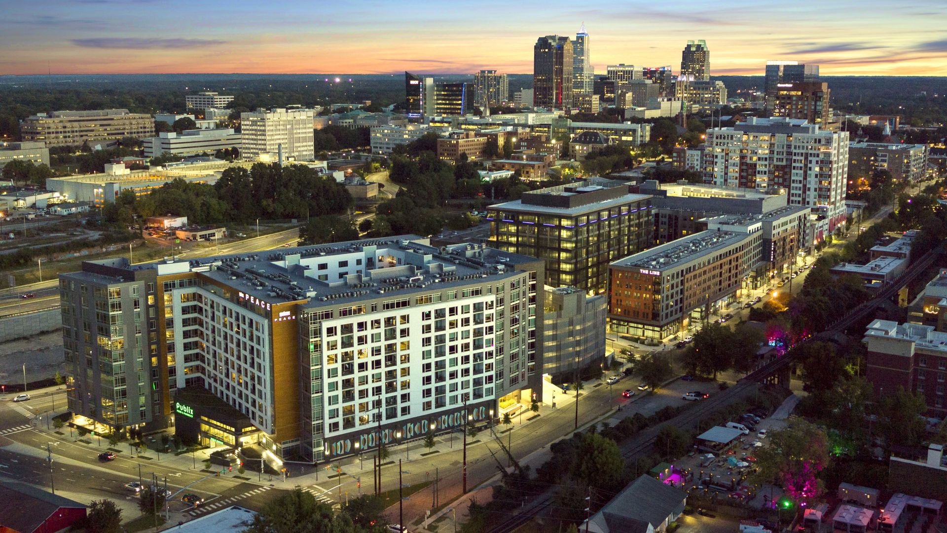 Aerial view of a modern cityscape at dusk with a clear sky, illuminated buildings, and streets, including a Publix and signs for Peace and The Line.