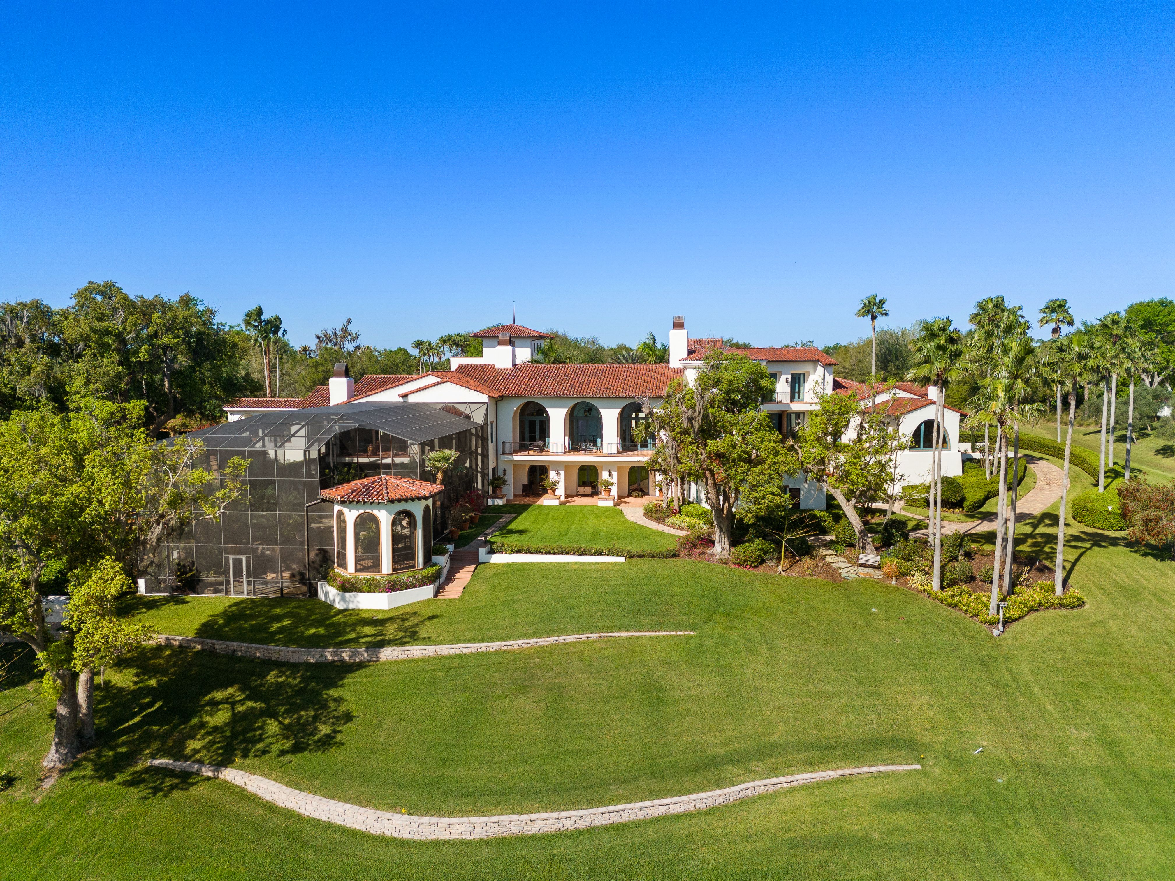 Large white Mediterranean-style house with red tiled roof, arched windows, and a screened pool area, surrounded by palm trees and green lawns under a clear blue sky.