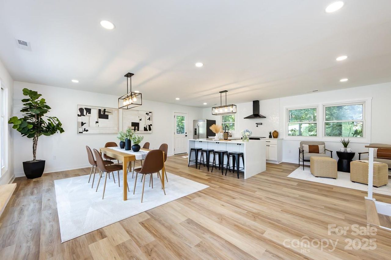 Bright open floor plan with white walls, light wood flooring, brown dining chairs around a wooden table, white kitchen island with black stools, and modern black light fixtures.