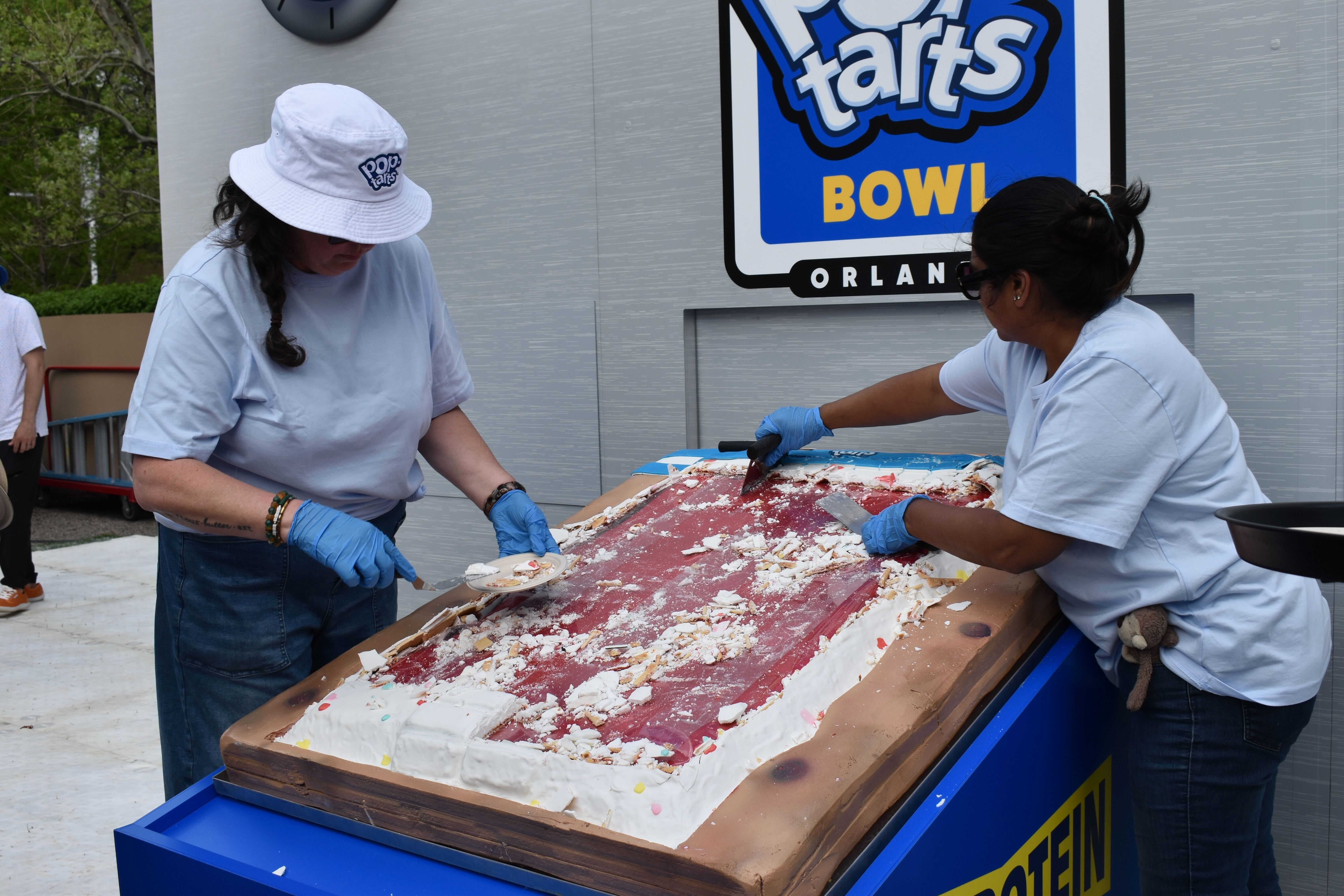Two women in light blue shirts and blue gloves cut a large, messy cake on a long serving surface outdoors; a white bucket hat and a blue sign reading BOWL appear in the background.
