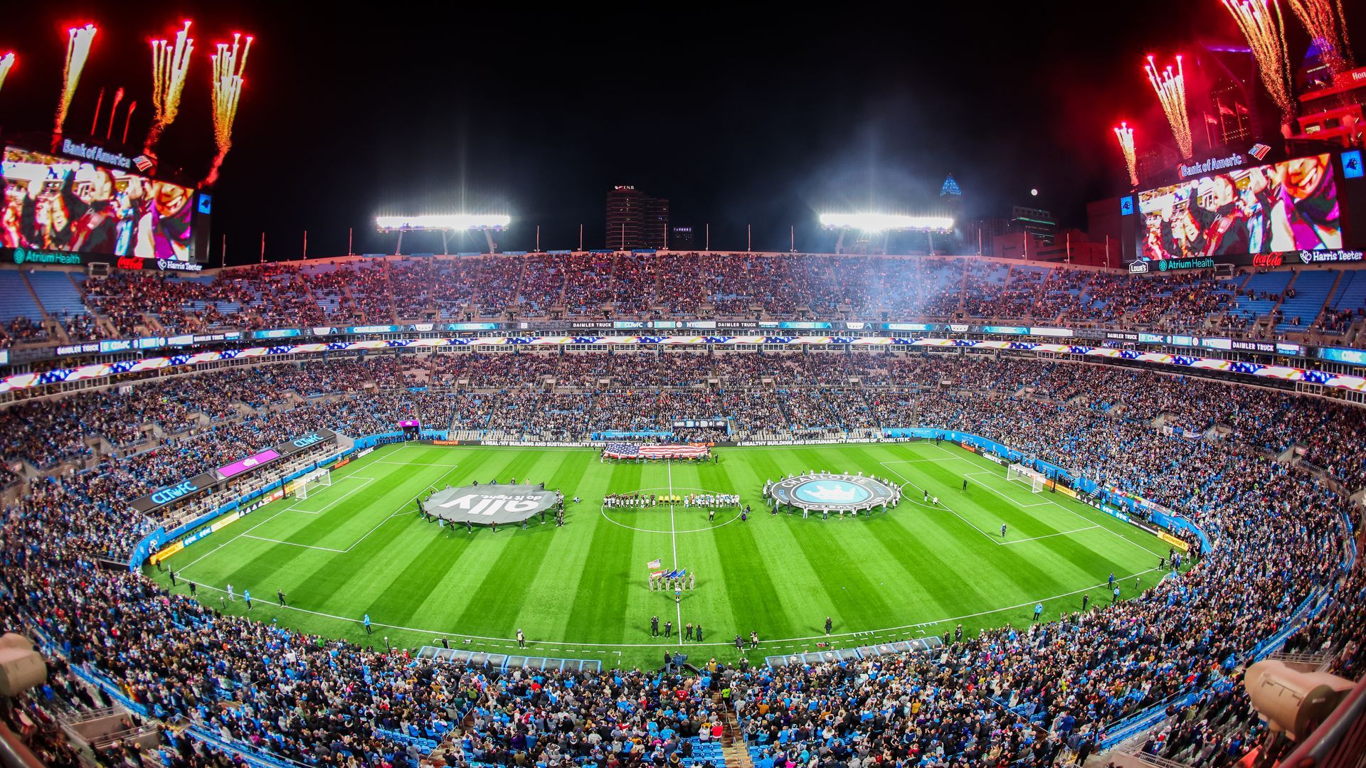 Fireworks at a Charlotte FC match at Bank of America Stadium in front of a packed house. Players and an American flag are on the field with the Charlotte skyline in the background. 