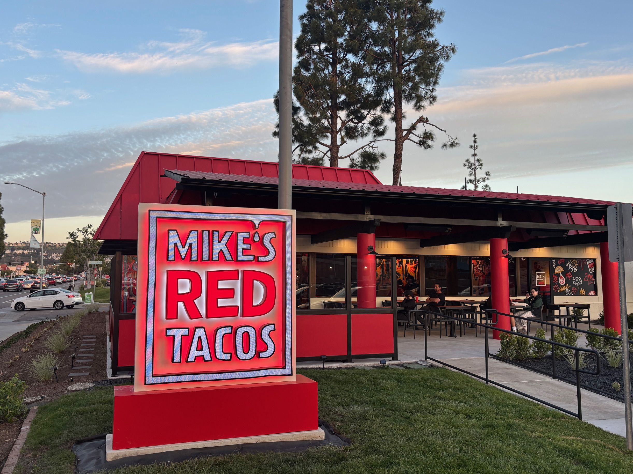 Exterior of a red-roofed restaurant with a large illuminated sign reading "MIKE'S RED TACOS". Outdoor seating under a covered patio with red columns, a ramp, diners at tables, trees and a blue sky.