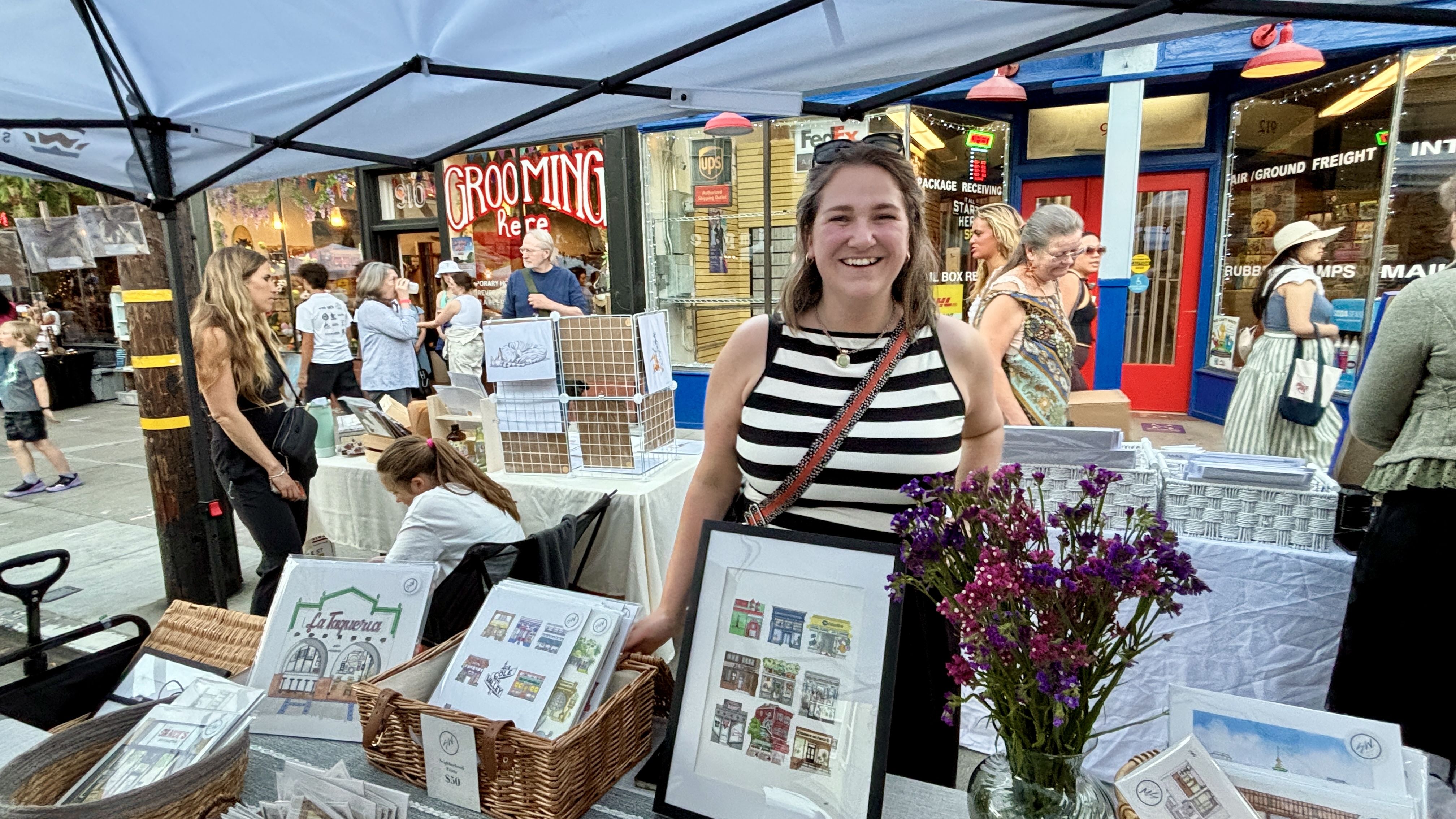 Smiling woman in a black-and-white striped sleeveless top stands at a market stall under a blue canopy, with framed prints, baskets, and purple flowers; shoppers and storefronts in the background.