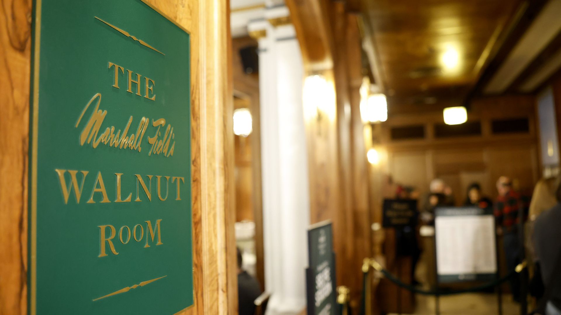 Green and gold sign reading "The Marshall Field's Walnut Room" mounted on a wood-paneled wall with a warmly lit, wood-toned interior and people blurred in the background.