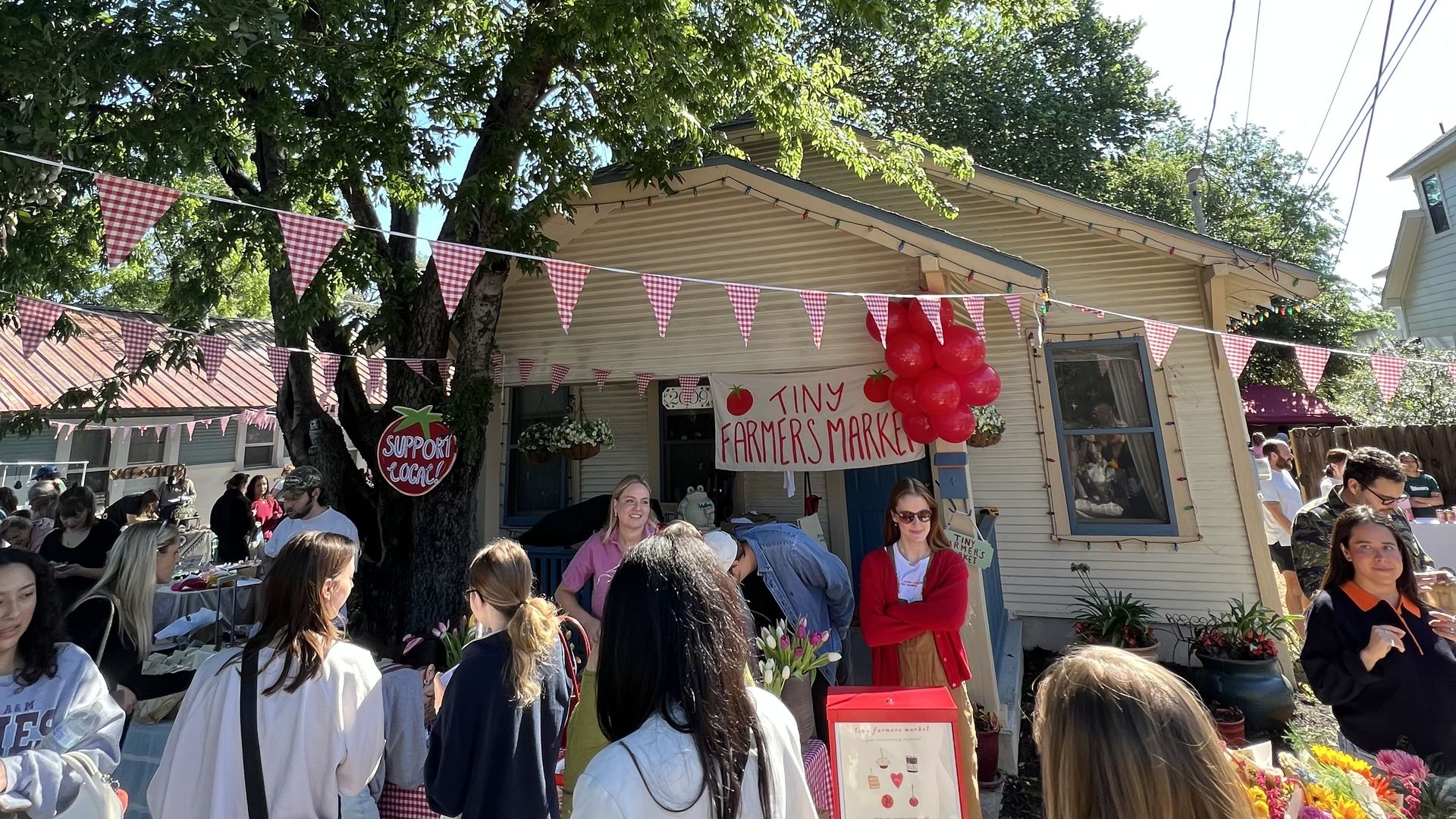 Outdoor farmers market under leafy trees with red-and-white bunting and string lights. A beige stall displays "Tiny Farmers Market" sign; red balloons and people browse flowers and produce.