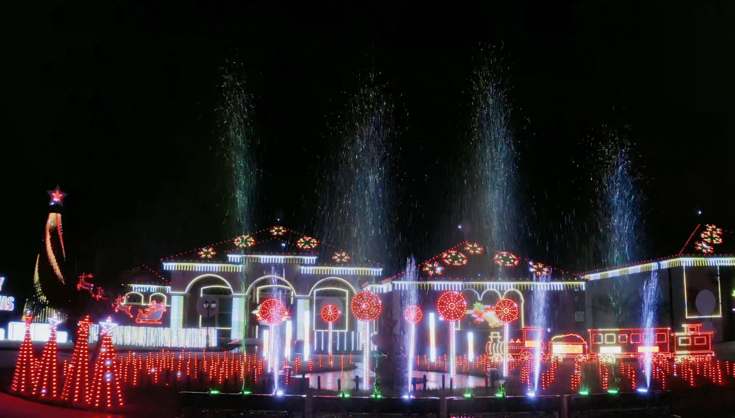 A front yard and home shown at night decked out in red, white and green Christmas lights along with several water fountains spraying high into the air.