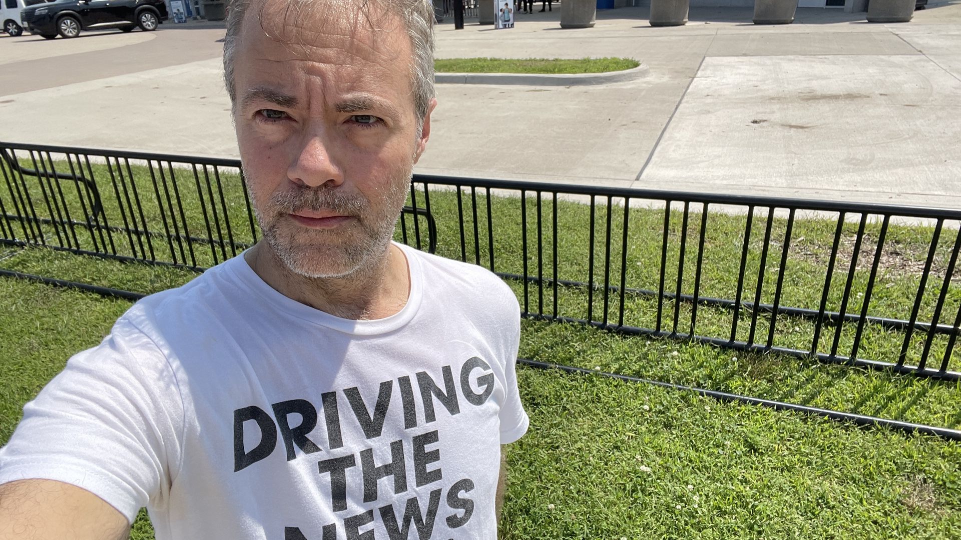 Selfie of a man with gray stubble wearing a white T-shirt that reads DRIVING THE NEWS, outdoors beside a black metal fence with green grass and a concrete plaza in the background.