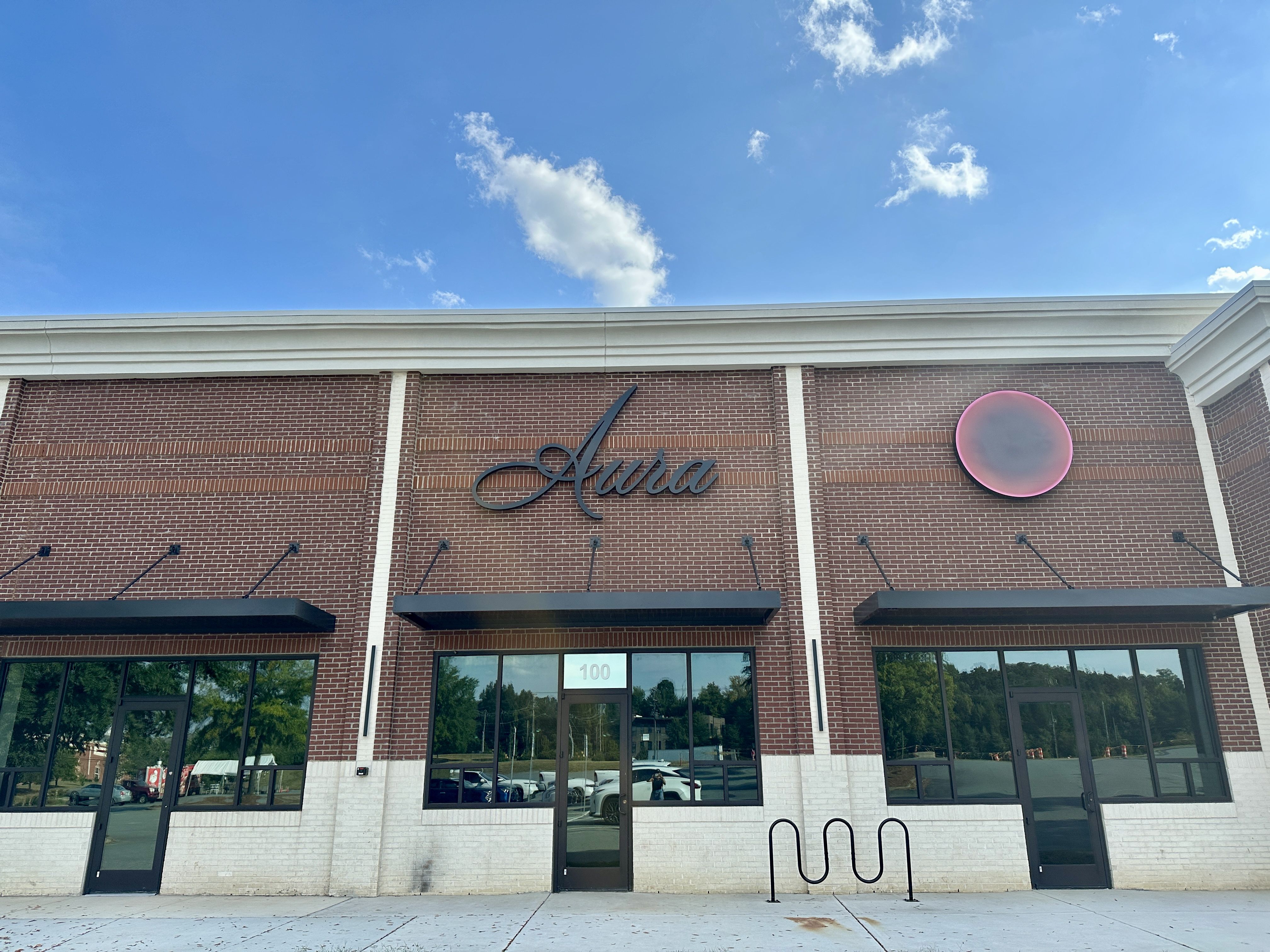 Brick building with white lower half and black doors and windows under black awnings. Sign reads "Aura" in script and a large round sign with pink border on right. Blue sky above.