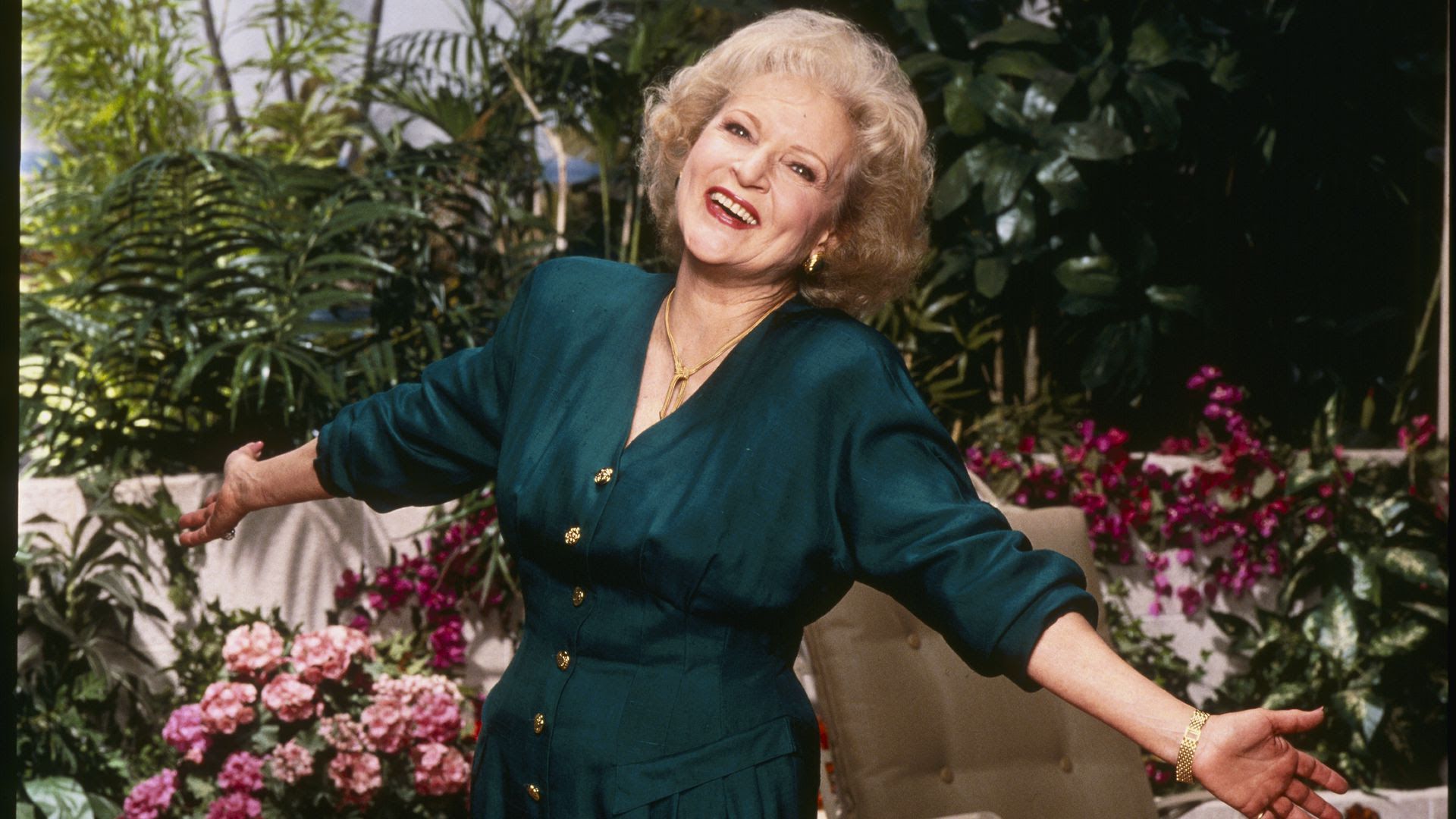 Betty White in a green dress in front of a flower garden.