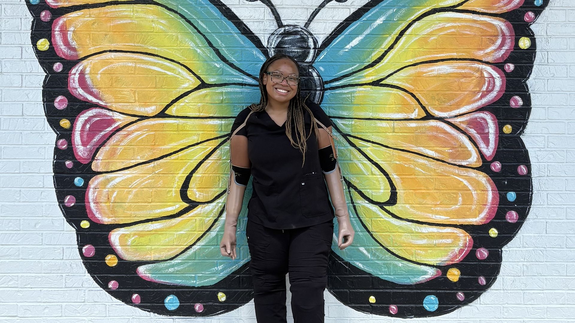 Smiling person in black stands in front of a colorful butterfly mural painted on a white brick wall with the words "Different Not Less" above it.