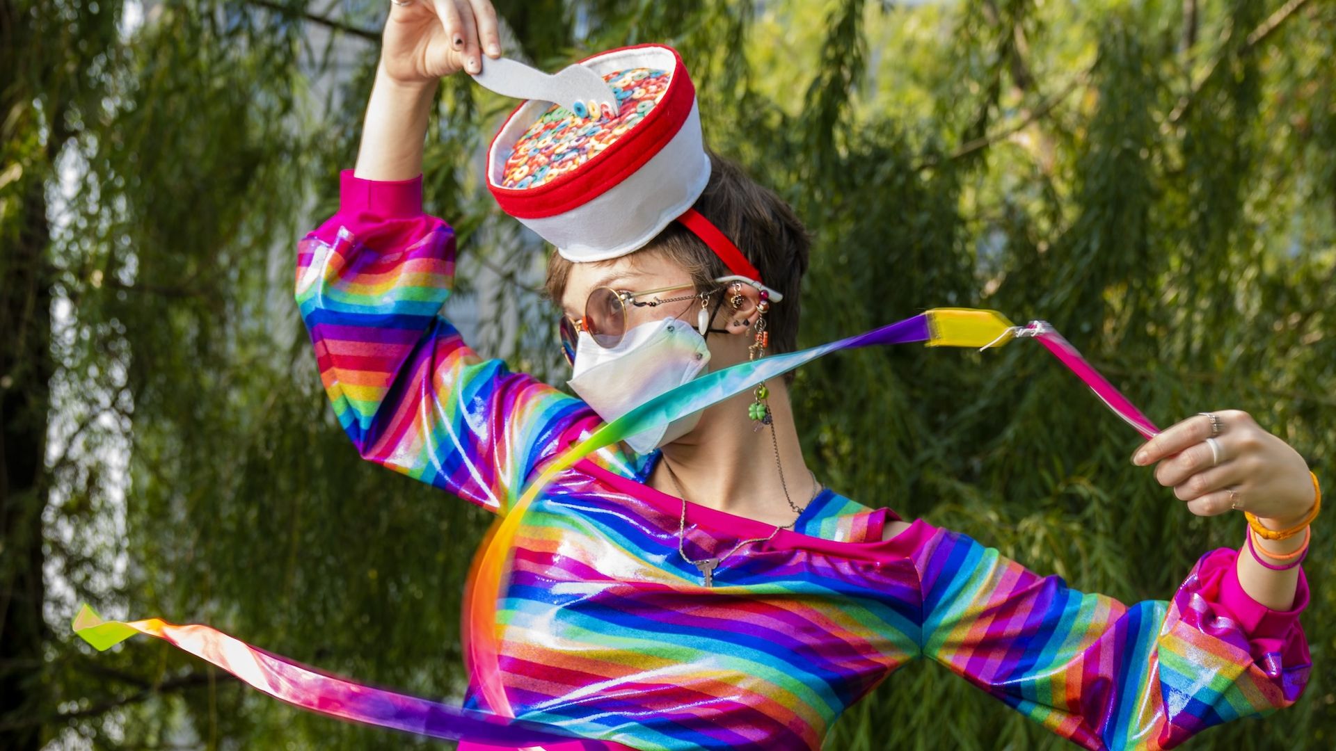 Person in a bright rainbow-striped outfit, wearing sunglasses and a face mask, tosses a red-and-white hat filled with colorful cereal while ribbons flutter in the breeze outdoors.