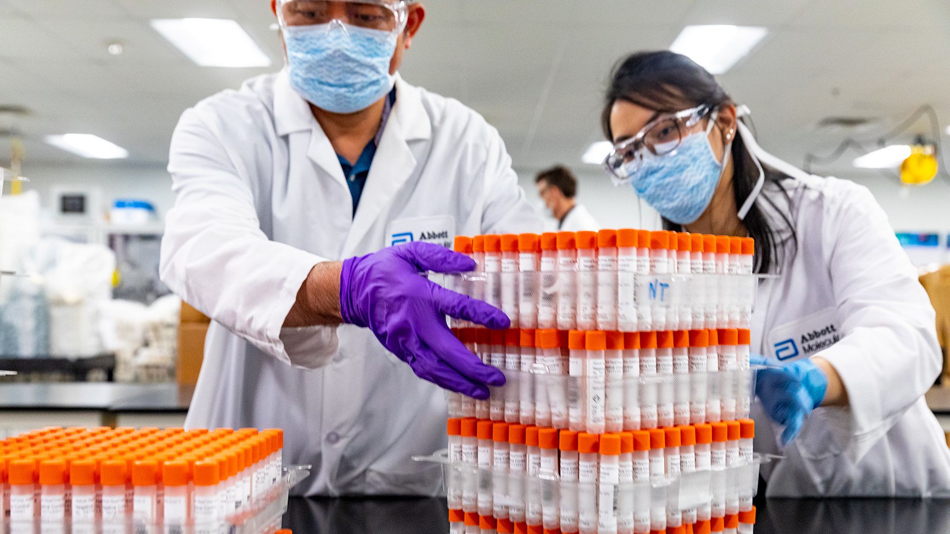 Two people in white lab coats handle stacks of test tubes with orange caps.