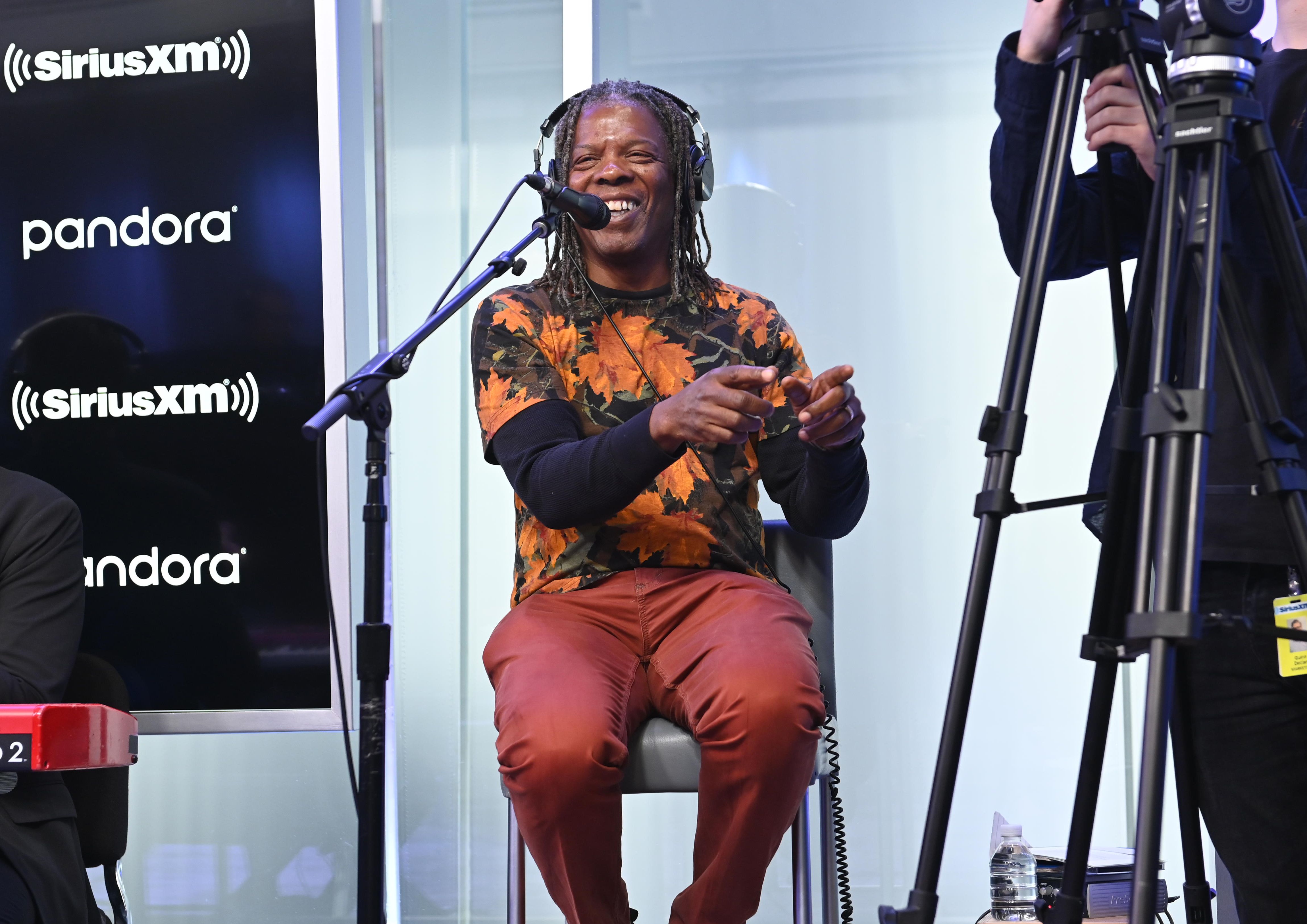 Smiling man with dreadlocks in an orange camouflage shirt and red pants sings into a microphone in a radio studio, wearing headphones. SiriusXM and Pandora logos appear on the left; camera gear on the right.