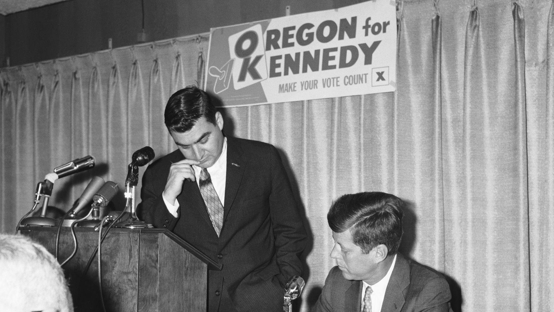 A sign reading Oregon for Kennedy hangs on a curtain behind a podium, with presidential candidate John F. Kennedy wearing a suit seated in front of it, writing on a pad as his press secretary Pierre Salinger, also in a suit, stands at a podium with microphones and looks at what Kennedy is writing. 