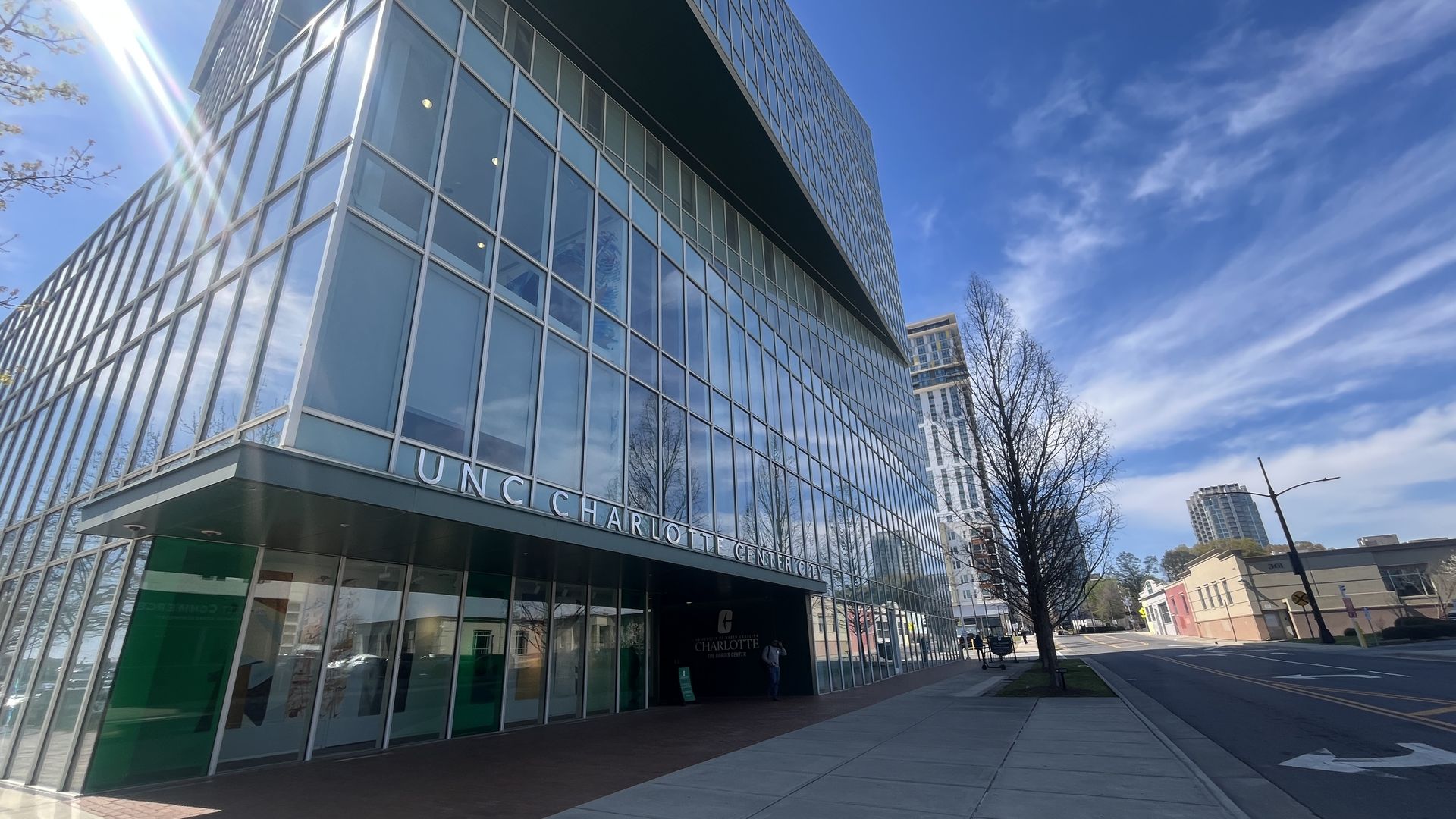 A modern glass-building with UNC Charlotte Center City sign, beside a sunlit sidewalk. Blue sky with clouds, lens flare, leafless trees, and a street with distant buildings.