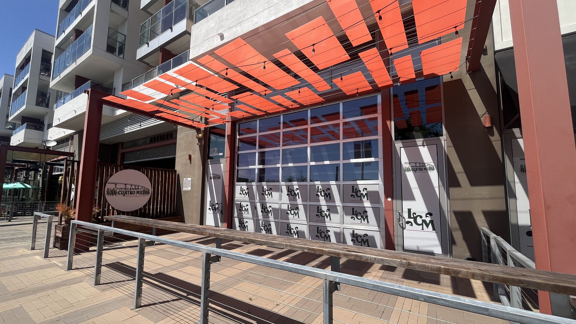 Modern multi‑story building with glass balconies and a bold orange canopy over the storefront. String lights hang above a patio with roll-up garage door windows and Las Cuatro Milpas logos. 