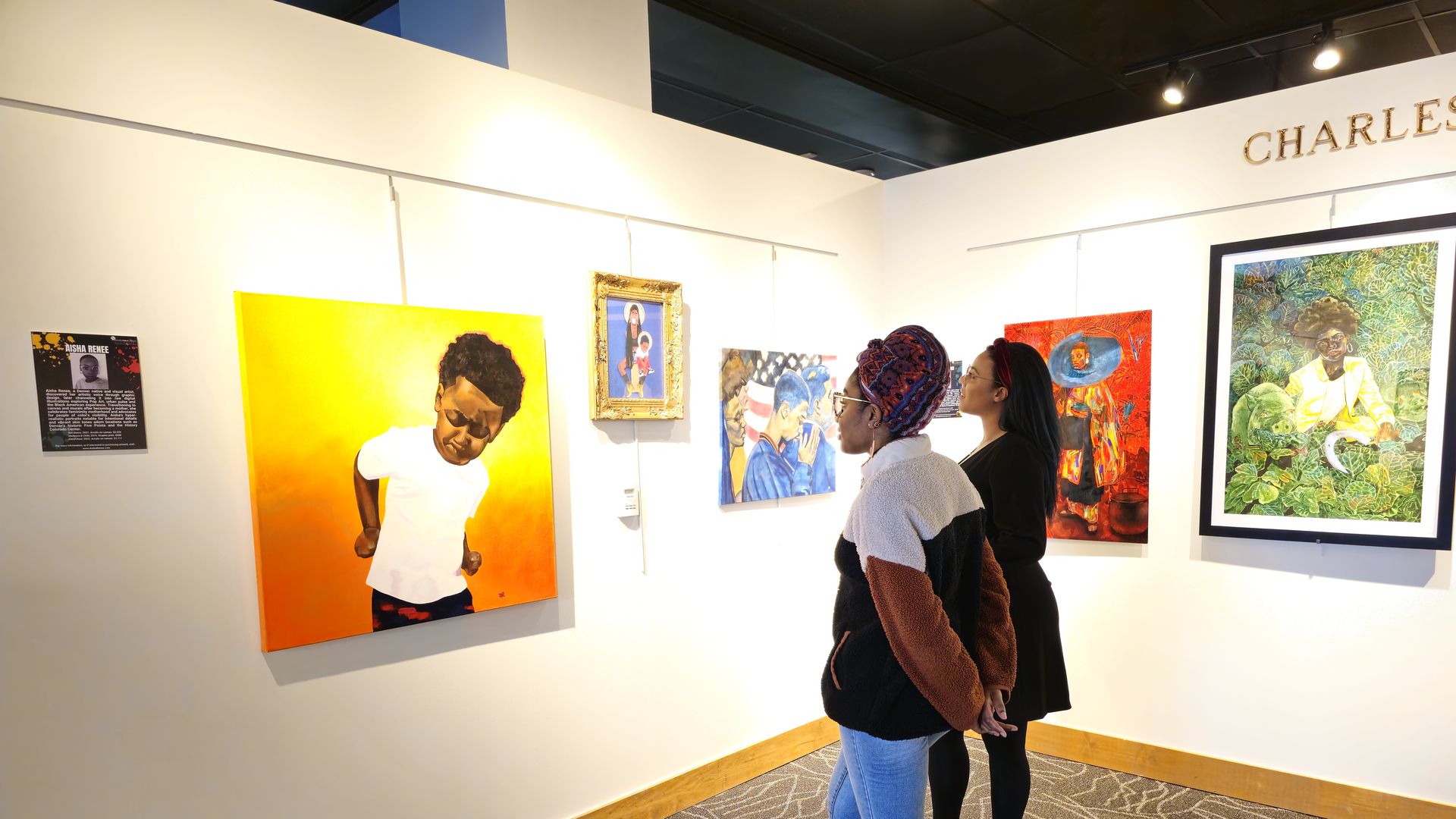 Two women look at a painting inside a gallery, with the painting on the left-hand side featuring a boy in a white t-shirt and an orange and yellow background. 