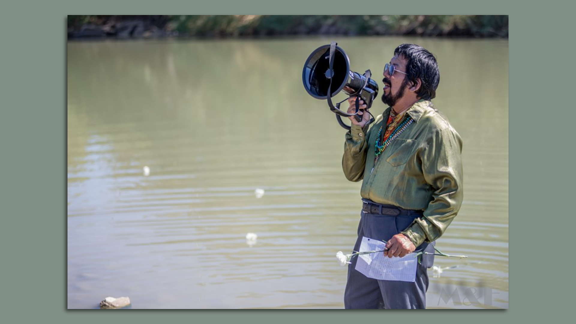 Nephtalí De León, wearing a green button down shirt and beaded necklaces, speaks into a megaphone in front of a river, while holding a flower and a sheet of paper.