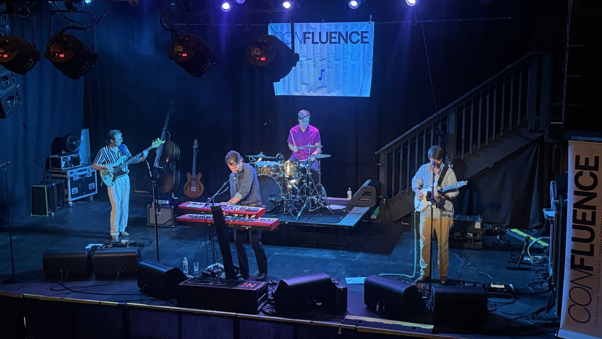 Four musicians perform on a dimly lit stage with blue and purple lighting. Instruments include keyboard, drums, bass guitar, and electric guitar. A banner behind reads "CONFLUENCE."