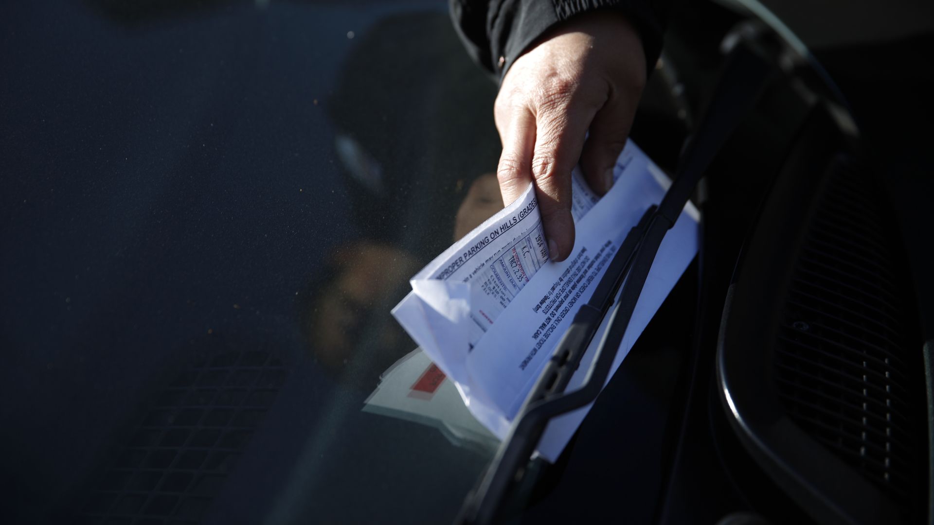 Photo of a hand slipping a parking citation paper behind a windshield wiper of a car
