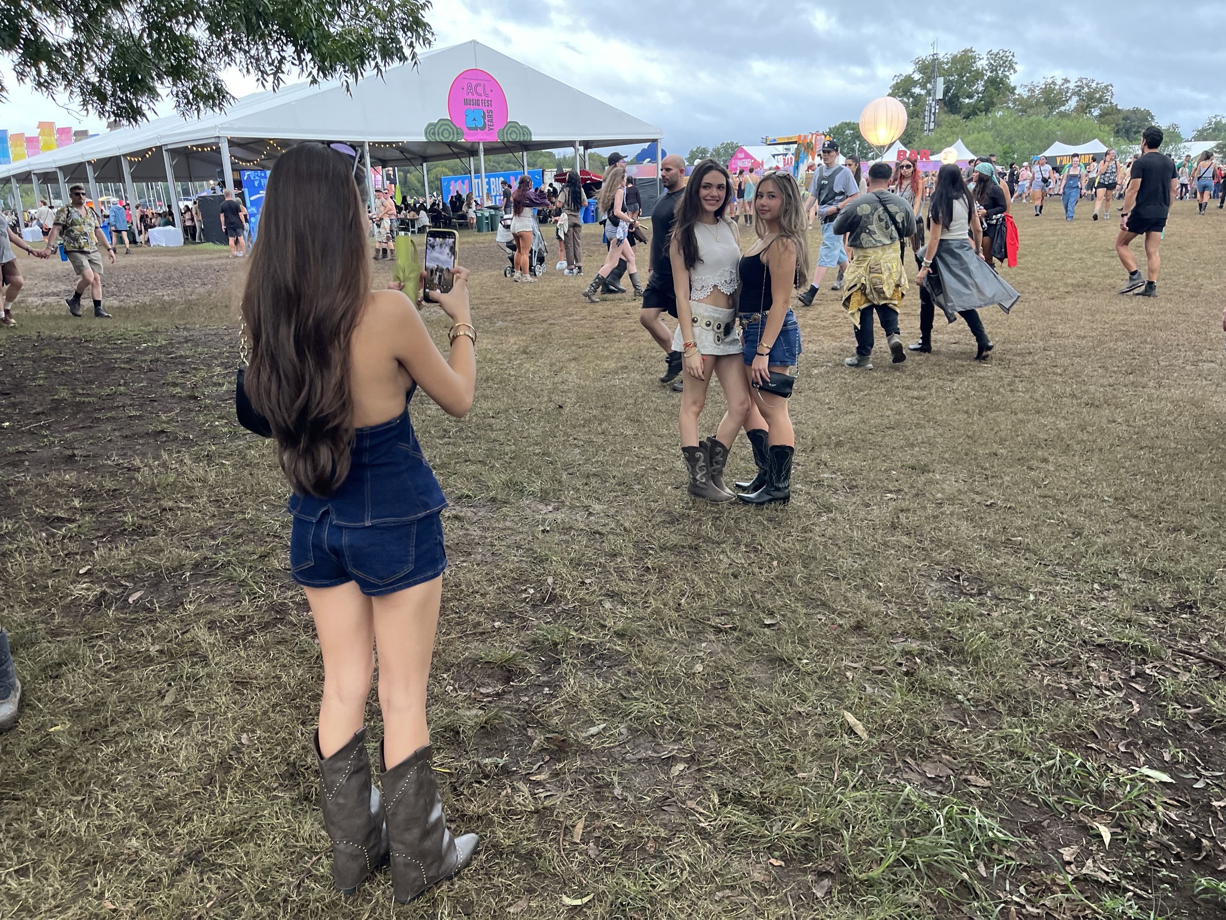 Crowd at outdoor festival with white tents and colorful banners. In foreground a woman takes a photo of two smiling girls in festival outfits, wearing boots, on a grassy field.