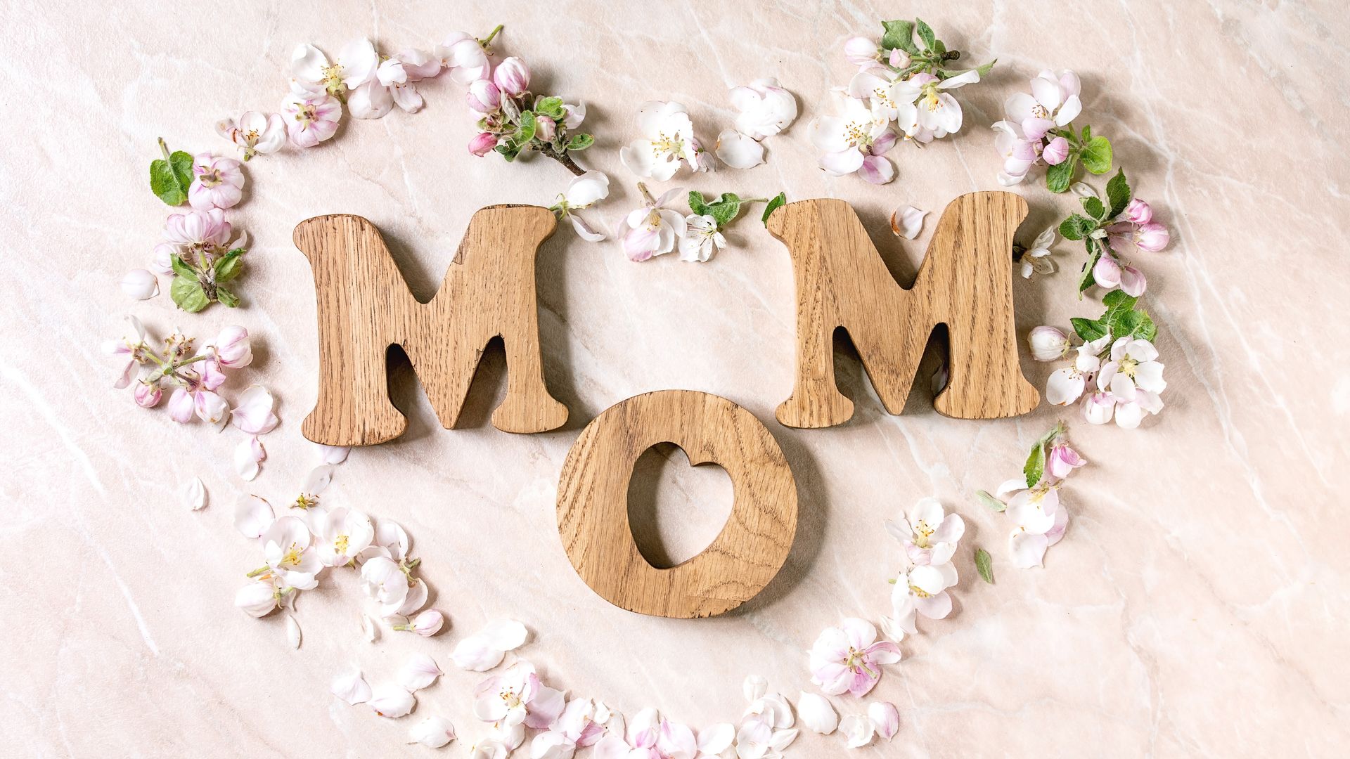 Wooden letters spell MOM with a heart-shaped cutout in the O, set on a pale marble background and surrounded by pink and white blossoms.