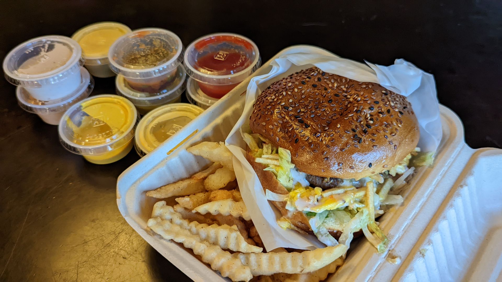 A hamburger sits with fries and plastic jars of sauce.