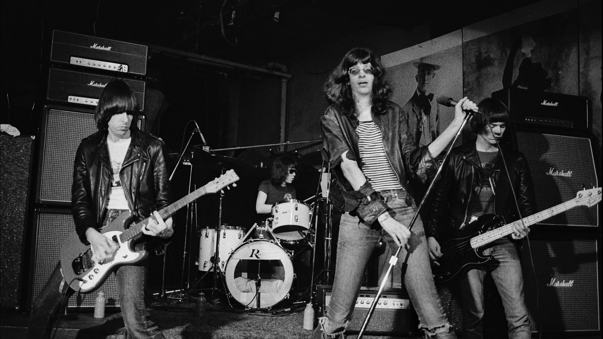 Black-and-white photo of a rock band performing on stage: lead singer in a striped shirt center, two guitarists beside him, drummer behind, Marshall amplifiers on both sides.