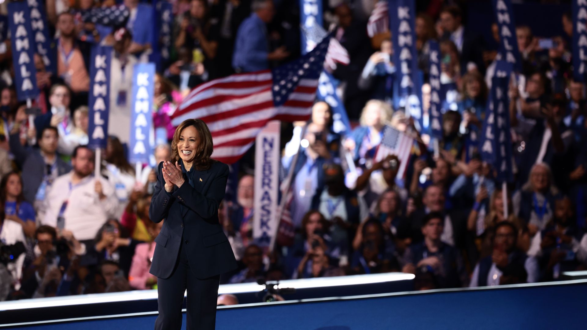 Vice President Kamala Harris, wearing a dark blue suit, standing on a blue stage in front of a crowd of DNC delegates holding blue "KAMALA" sign sticks. 