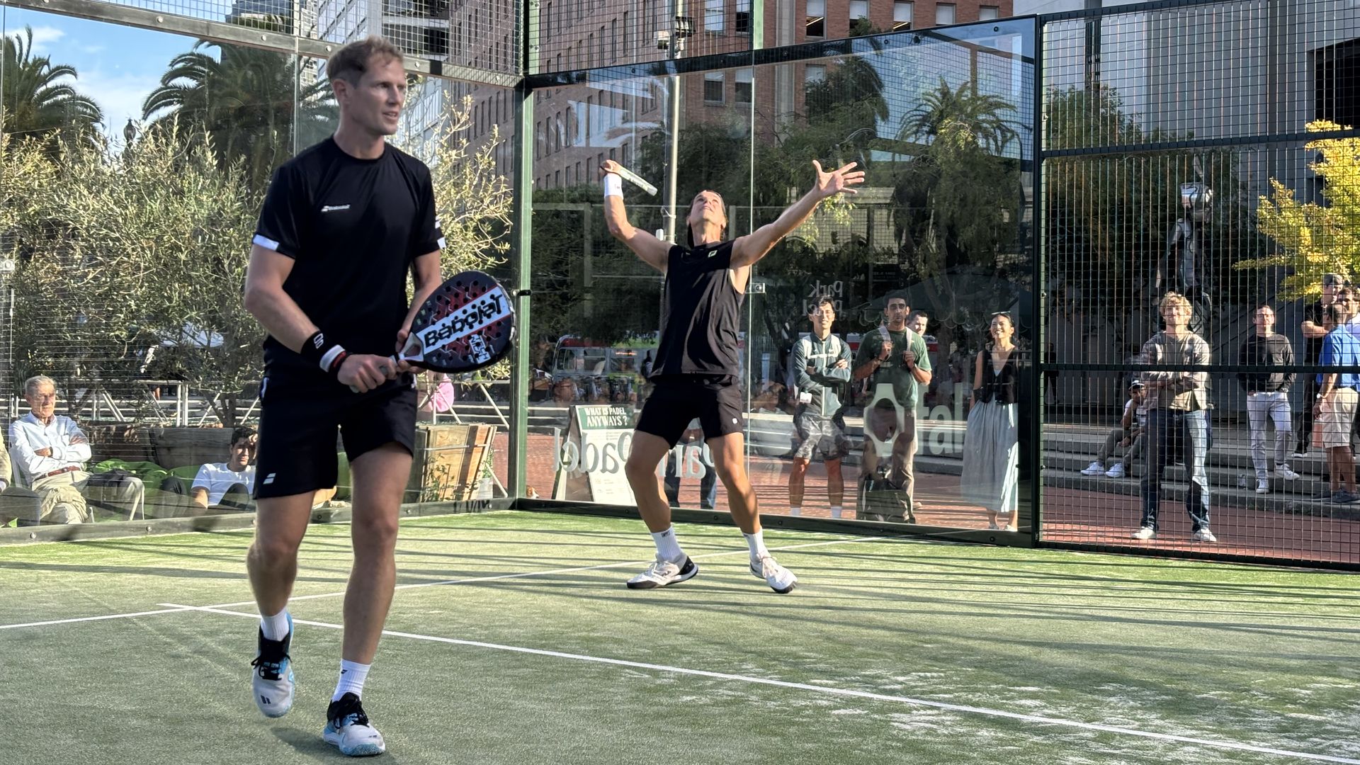 Two men playing padel on a glass court in an urban setting; one prepares to hit the ball while the other raises his paddle and arm to serve, spectators watch outside the court.