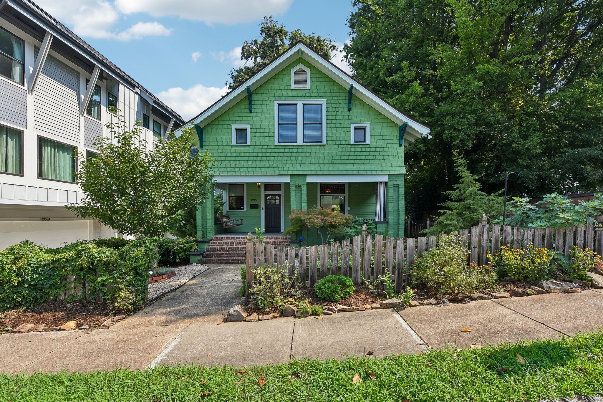A lime-green, two-story house with white trim and a steep gabled roof sits between a white townhouse on the left and dense green trees on the right. Brick steps lead to a front porch with a bench.