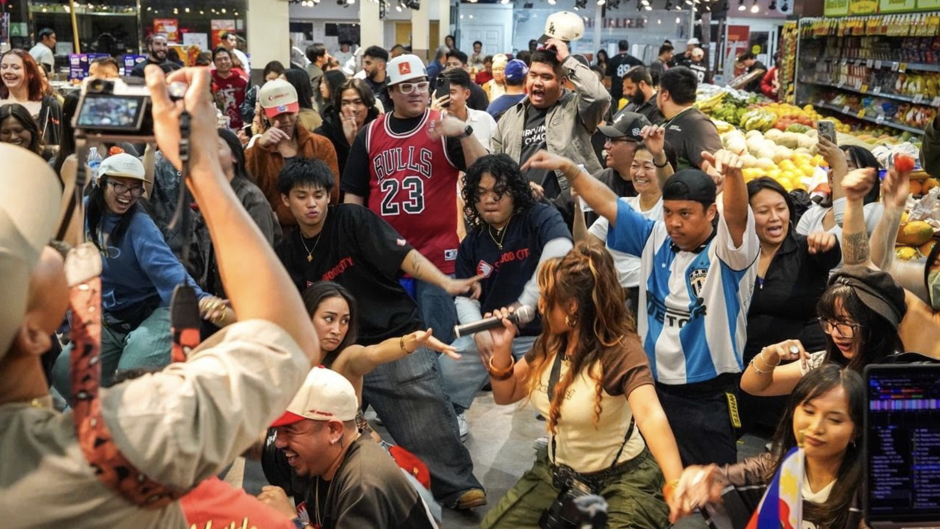Large crowd of shoppers dancing and filming in a busy supermarket, under bright overhead lights; many wear Bulls 23 jerseys, hats, and casual outfits, with fruit displays in the background.