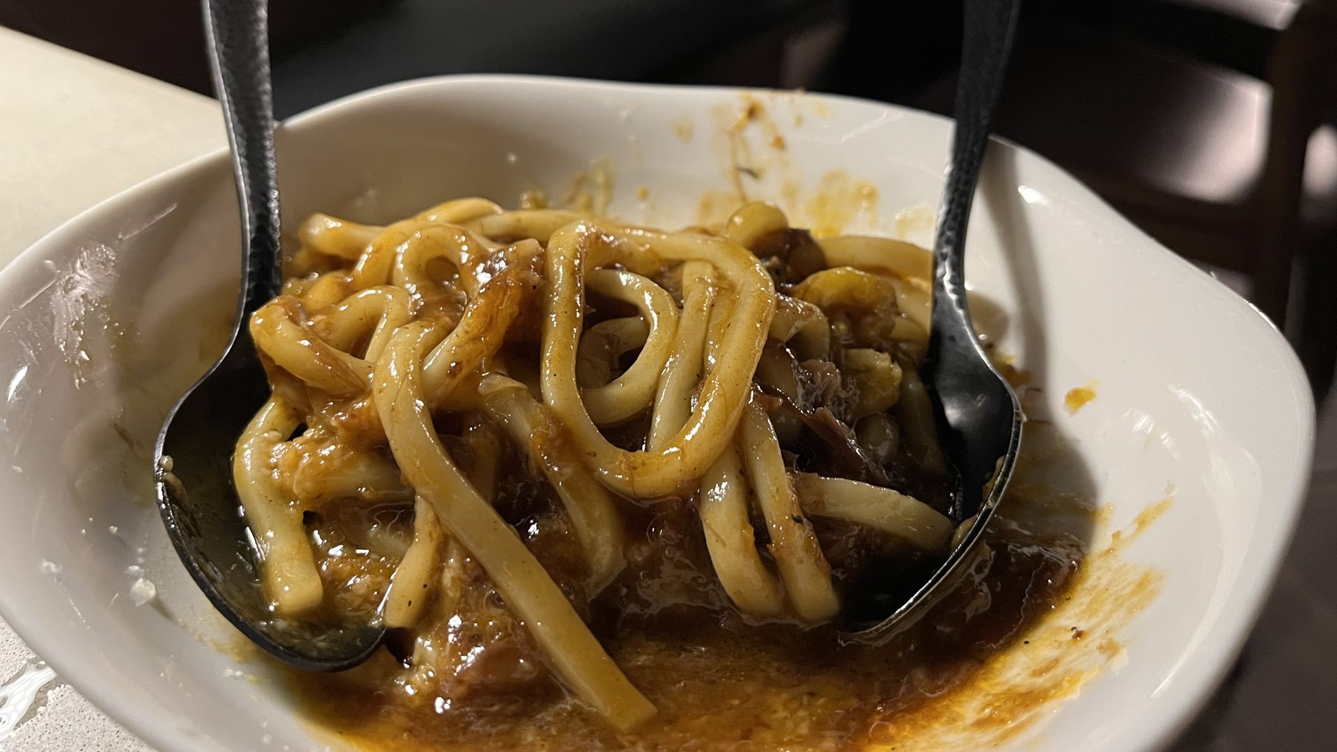 White bowl with thick noodles coated in a rich, brown sauce, two metal spoons placed inside the bowl on a white surface with a blurred background.