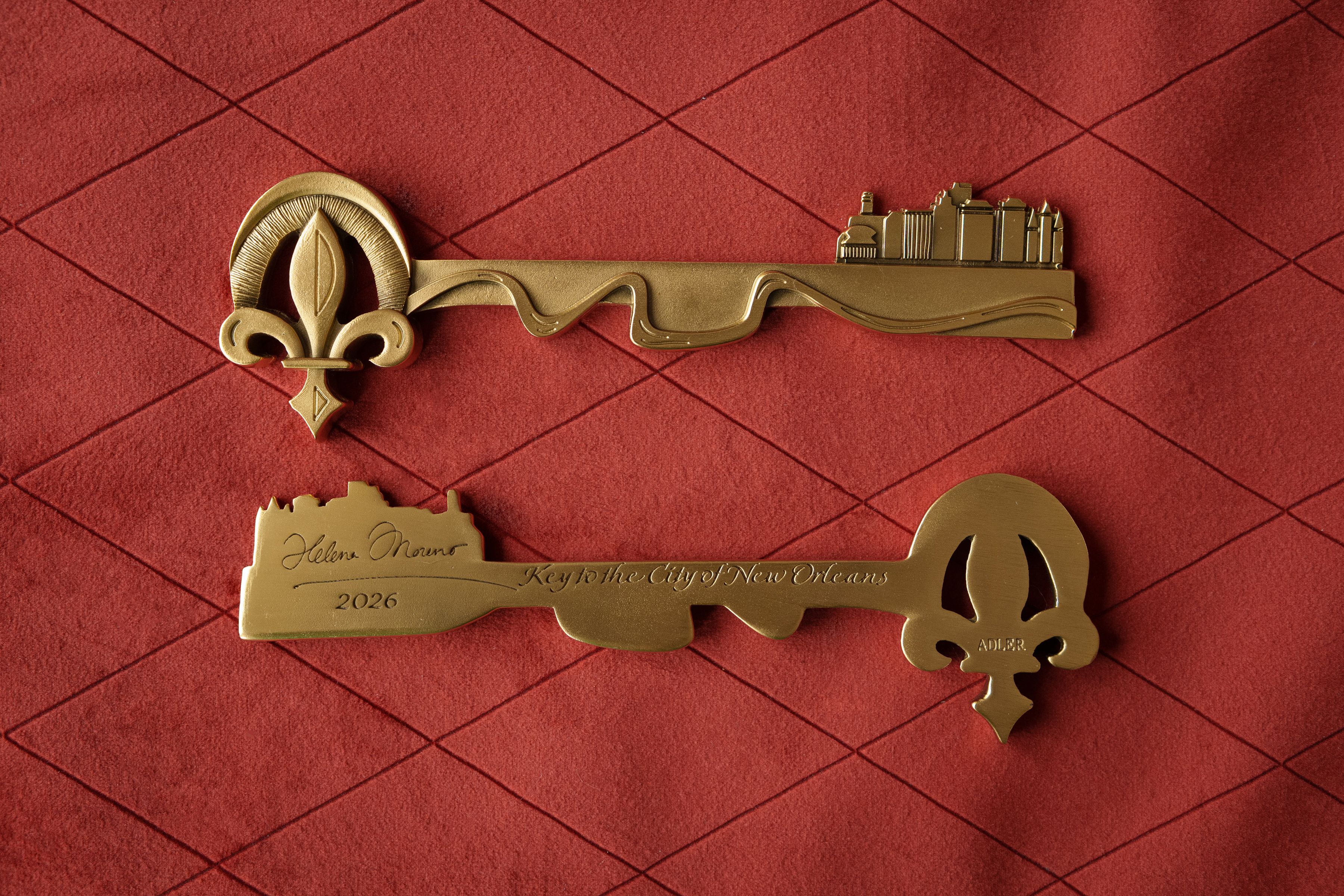 Gold ceremonial key with a fleur-de-lis handle and a city skyline on the shaft's right end, engraved "Key to the City of New Orleans" and "Helena Moreno 2026" on a red quilted background.