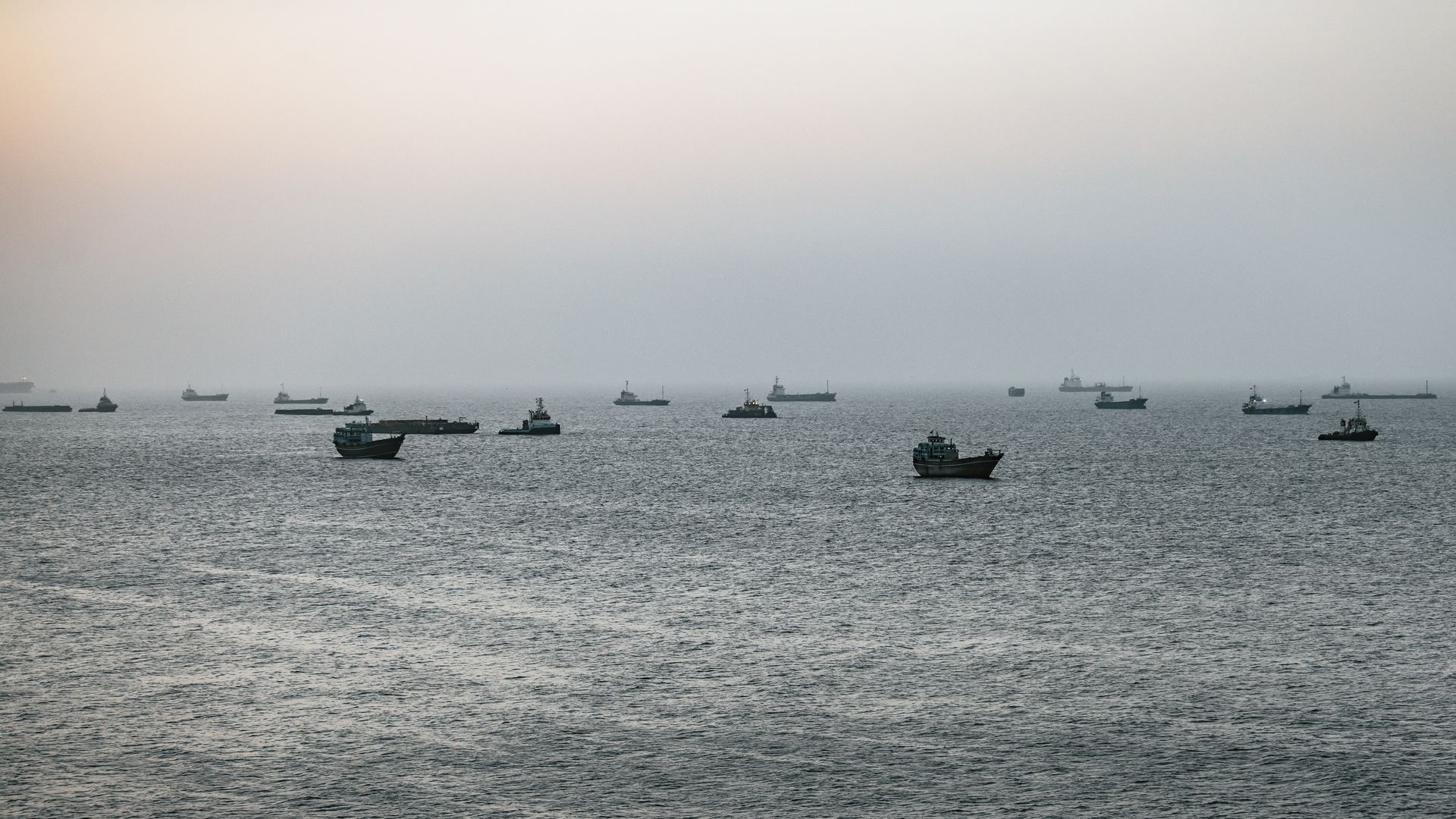 Many small to mid-sized ships spread across a calm sea, silhouetted against a pale pastel sunrise with soft peach, pink, and blue sky near the horizon.
