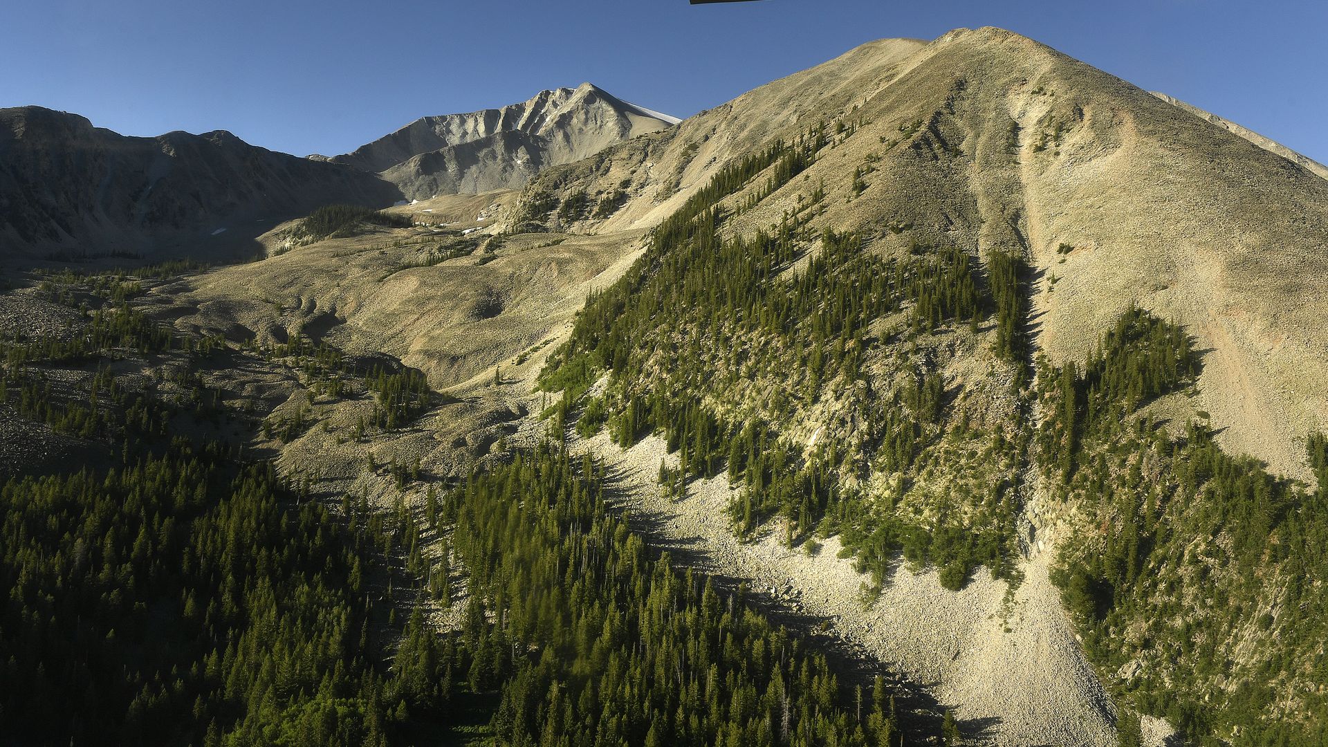 View of rocky, tree-dotted mountain slopes under a clear blue sky, taken from a small plane showing a part of its wing at the top of the image.