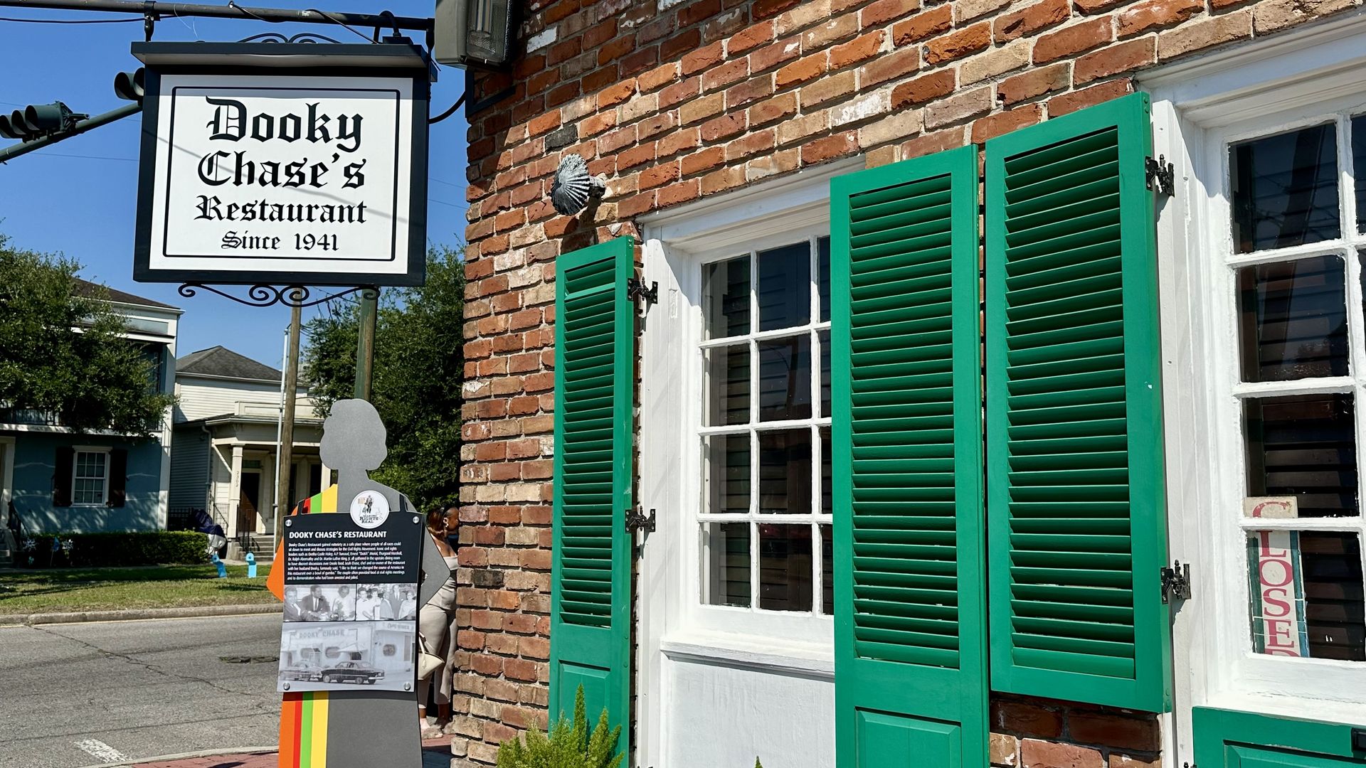 Exterior of Dooky Chase's Restaurant with a brick wall, green shutters, and a sign reading "Dooky Chase's Restaurant Since 1941" under a blue sky on a sunny day.
