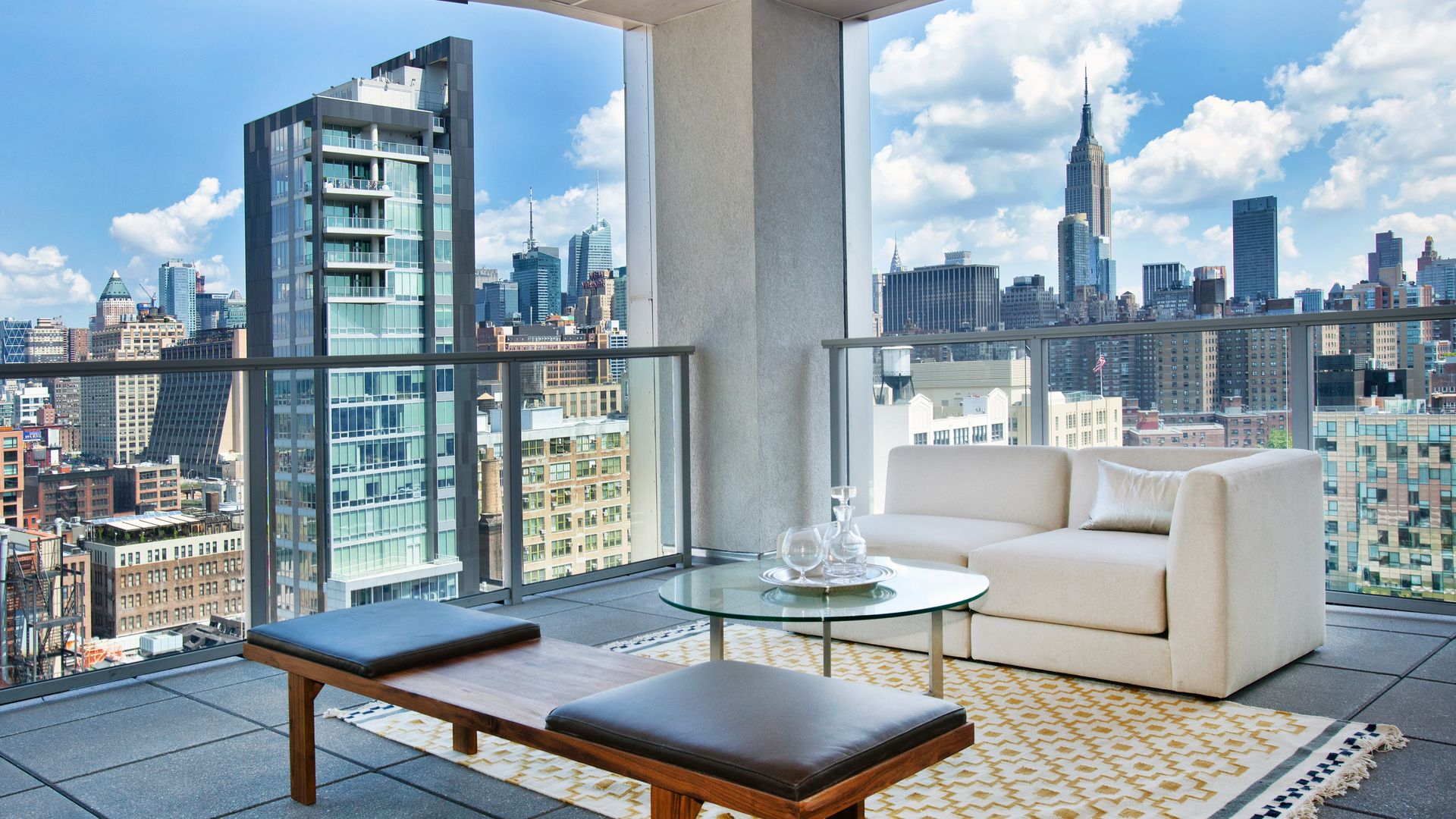 Modern balcony overlooking a city skyline. White outdoor seating around a glass coffee table on a yellow geometric rug, glass railing, and blue sky with fluffy clouds behind tall skyscrapers.