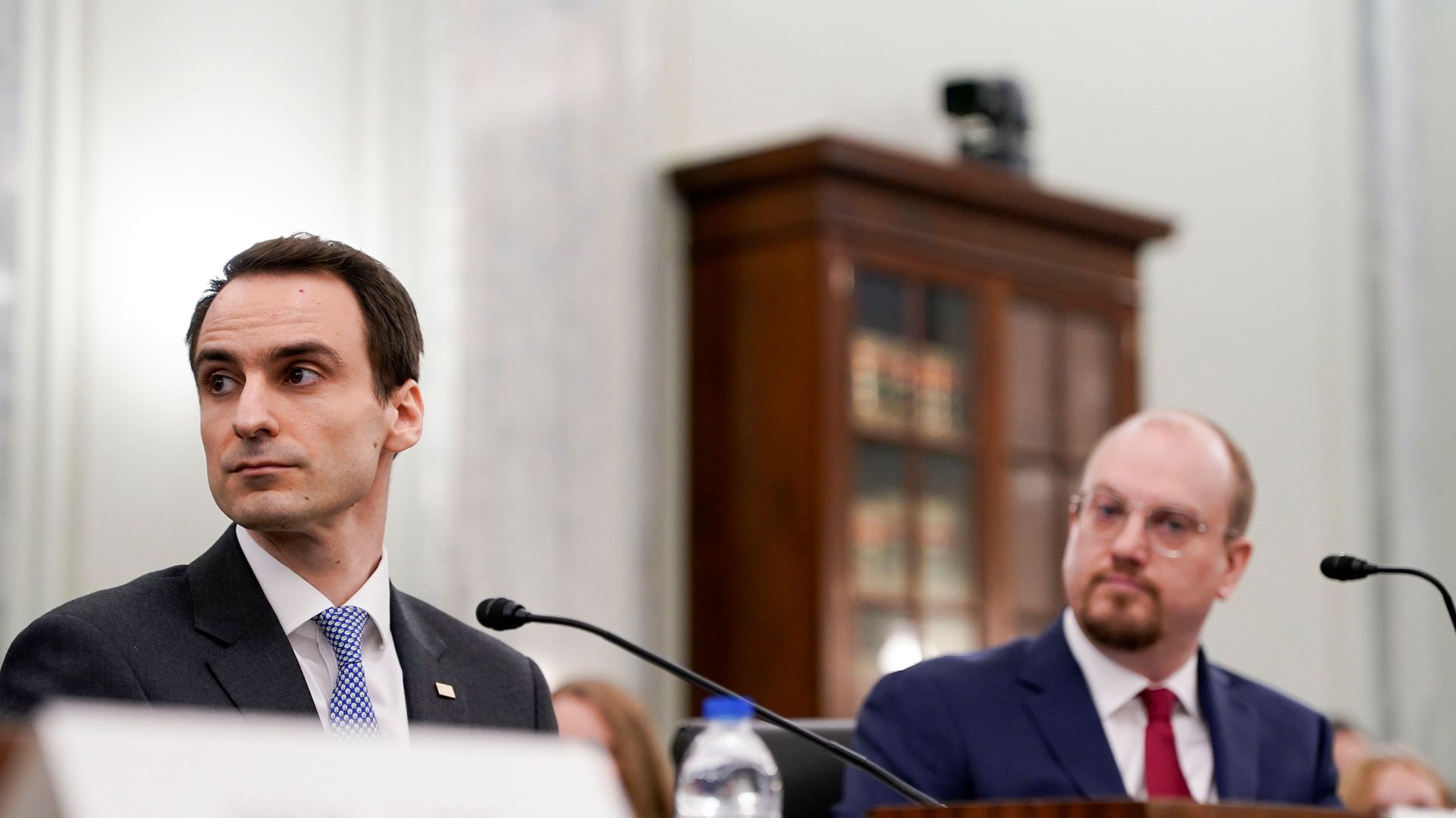 Two people in suits and ties sit in front of microphones
